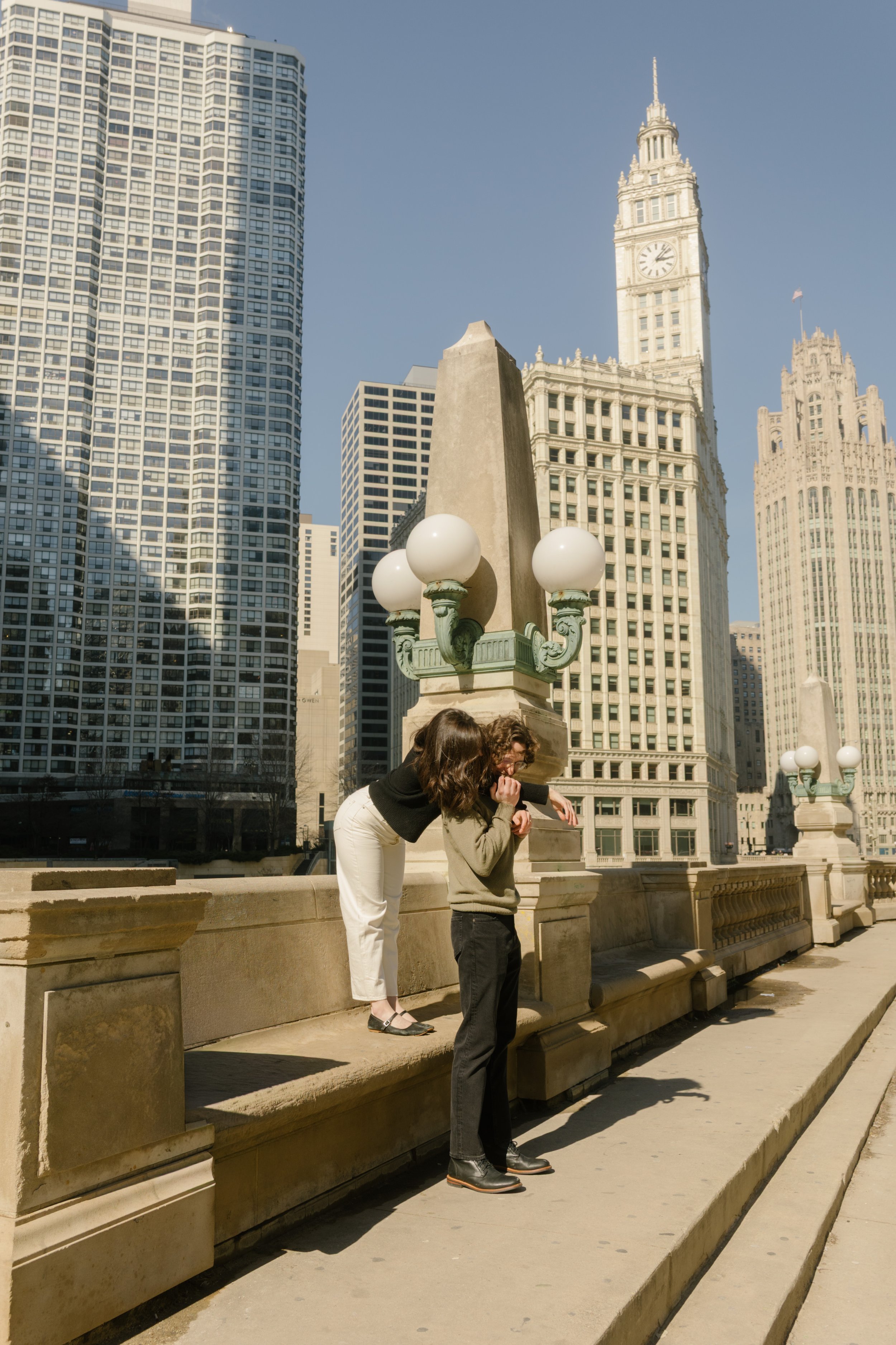 Playful moment of one partner leaning over the bridge railing while the other stands below, framed by tall Chicago buildings and bright afternoon light