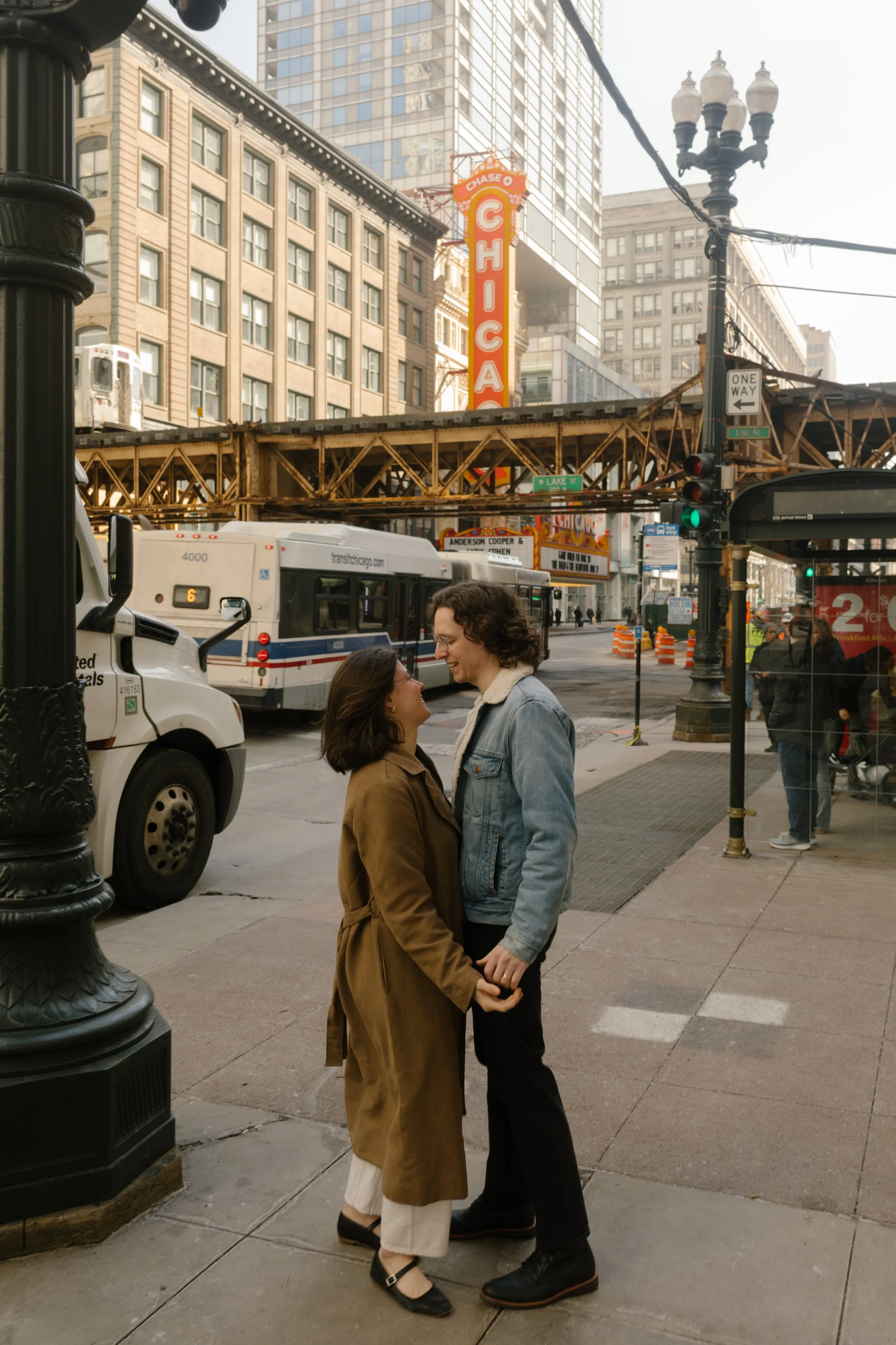 Couple holding hands and smiling at each other on a downtown Chicago street with the elevated train tracks and theater sign behind them