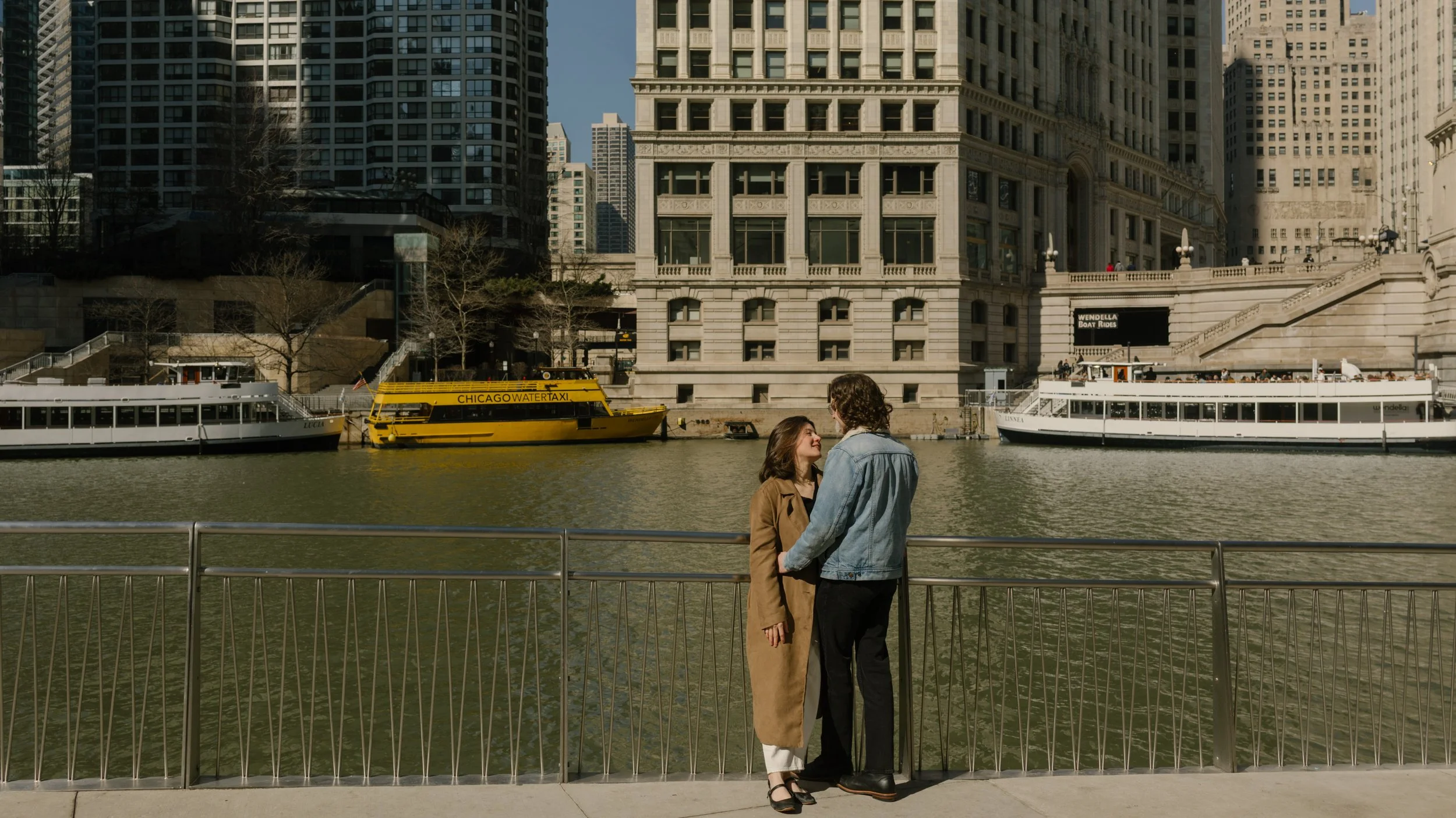 Couple standing together along the Chicago River with a yellow water taxi and city buildings in the background during engagement photos in Chicago