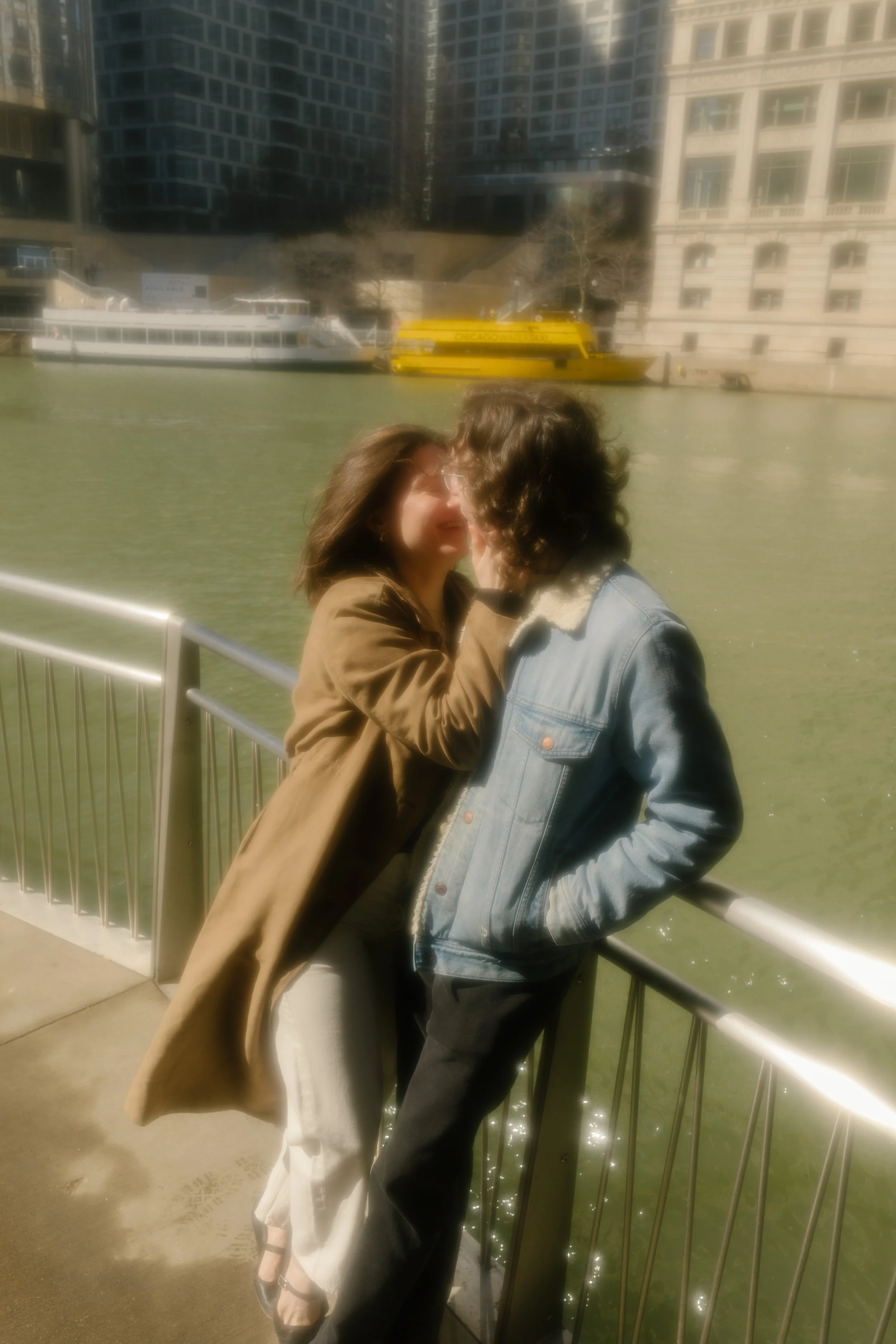 Soft-focus image of couple leaning against a riverwalk railing with water and passing boats behind them