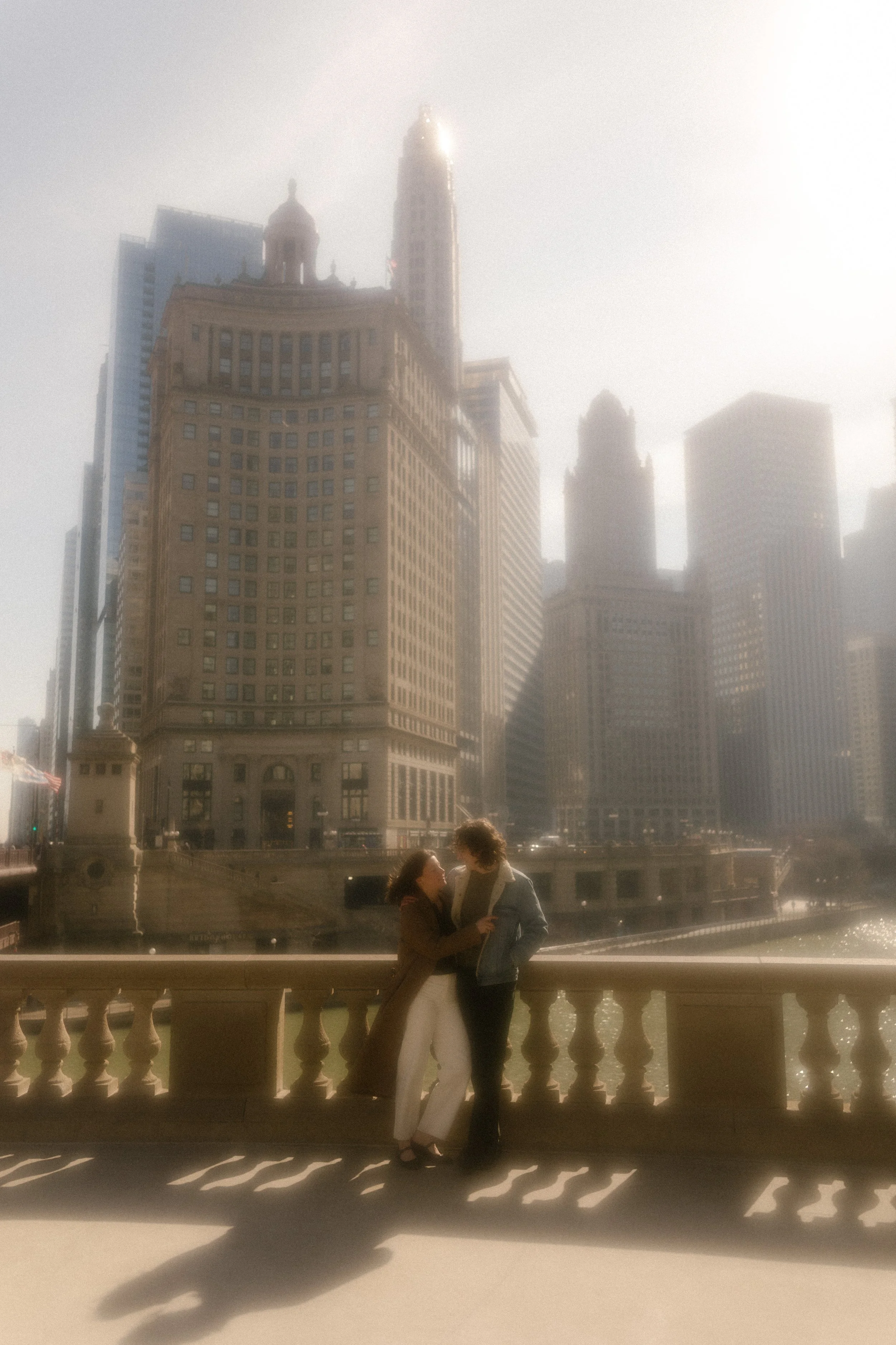 Wide, hazy shot of couple standing together on a bridge with Chicago skyline glowing in the background during engagement photos in Chicago