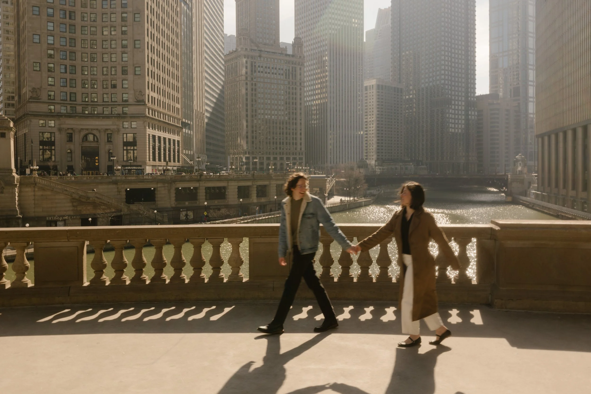 Couple holding hands and walking across a bridge over the Chicago River with skyline views during engagement photos in Chicago
