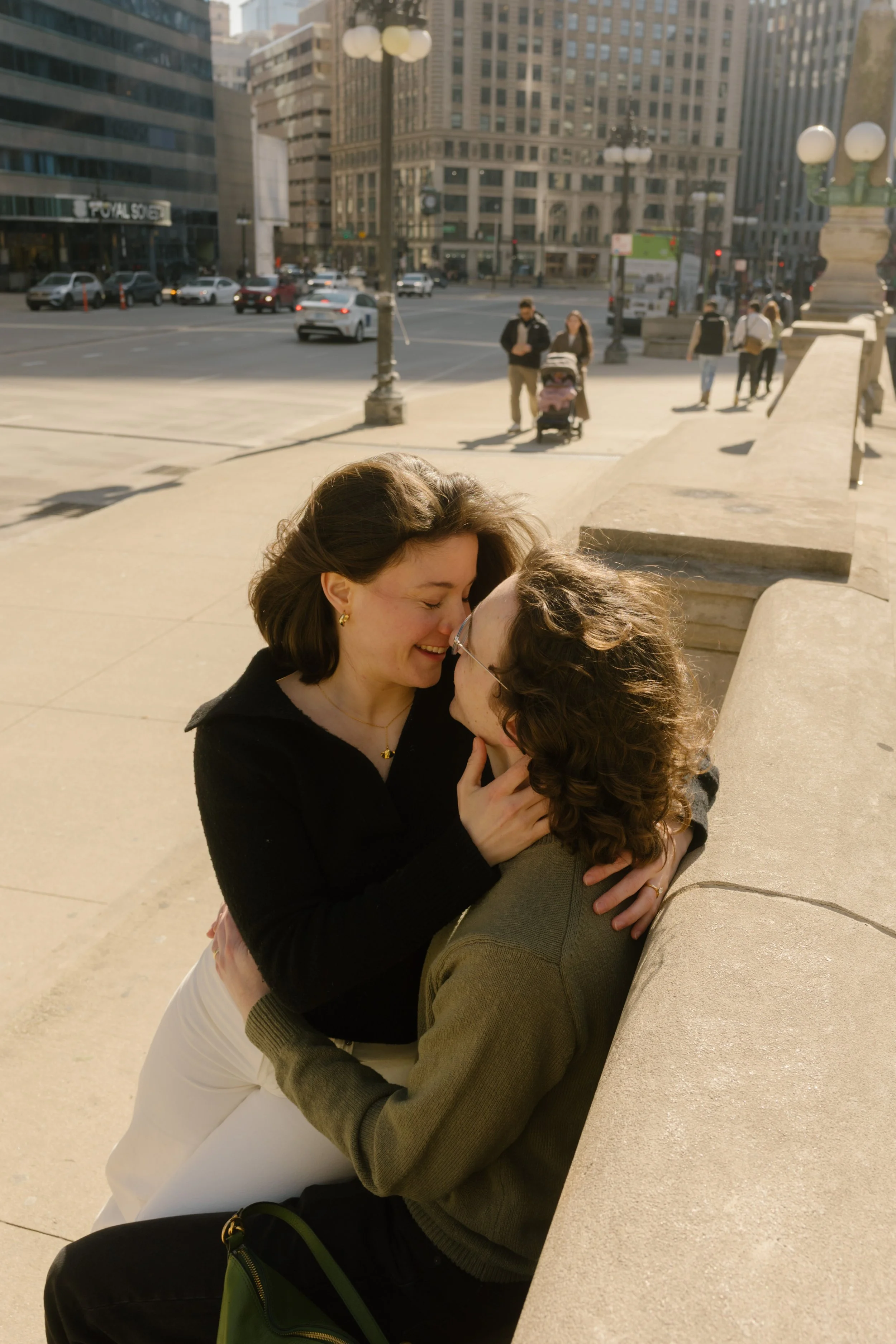 Intimate moment of couple sitting and laughing together along a city sidewalk with traffic and pedestrians in the background during engagement photos in Chicago