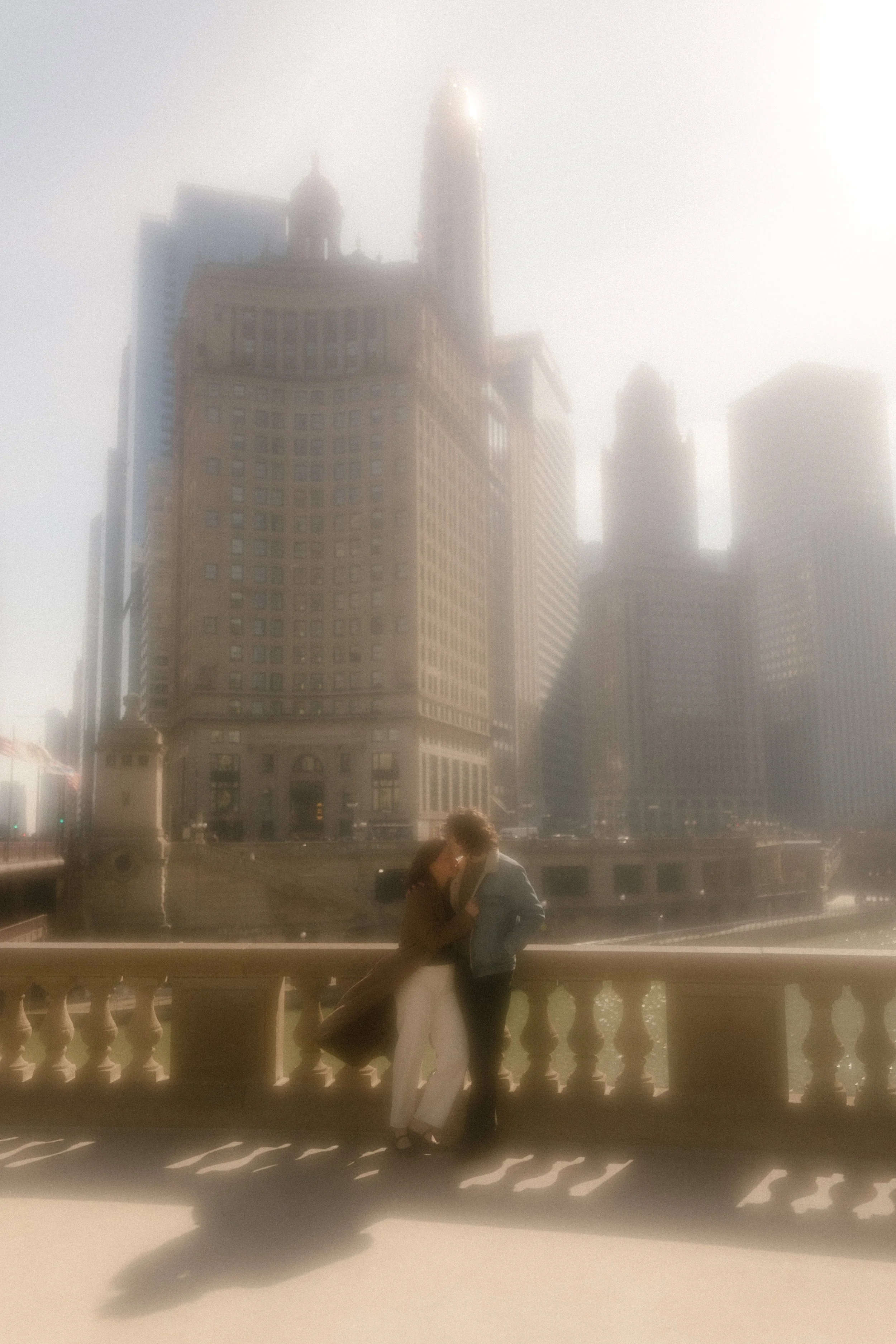 Soft, hazy photo of couple standing close together on a bridge with glowing light and city skyline during engagement photos in Chicago