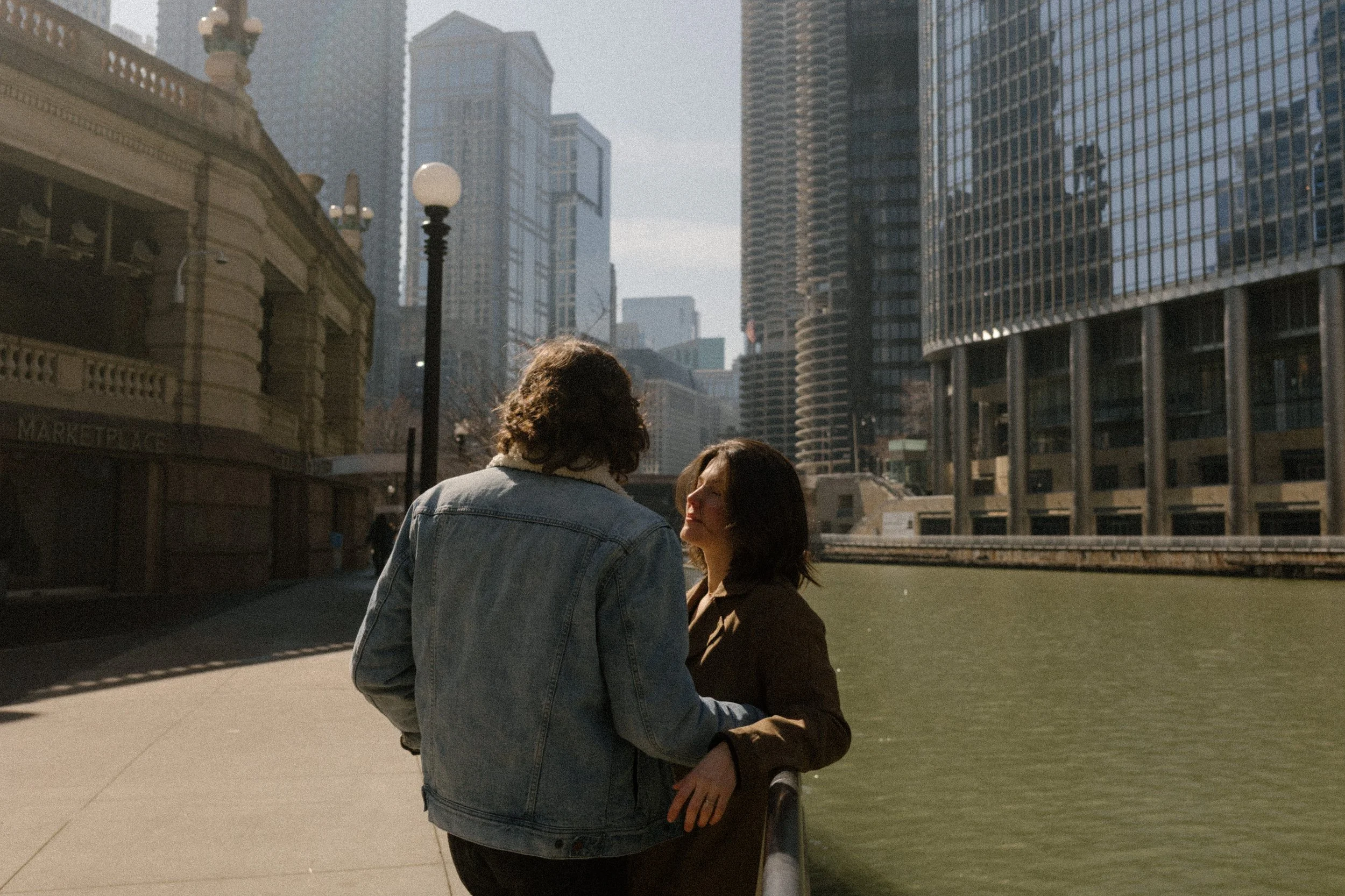 Couple leaning against the riverwalk railing, talking quietly with downtown buildings rising behind them