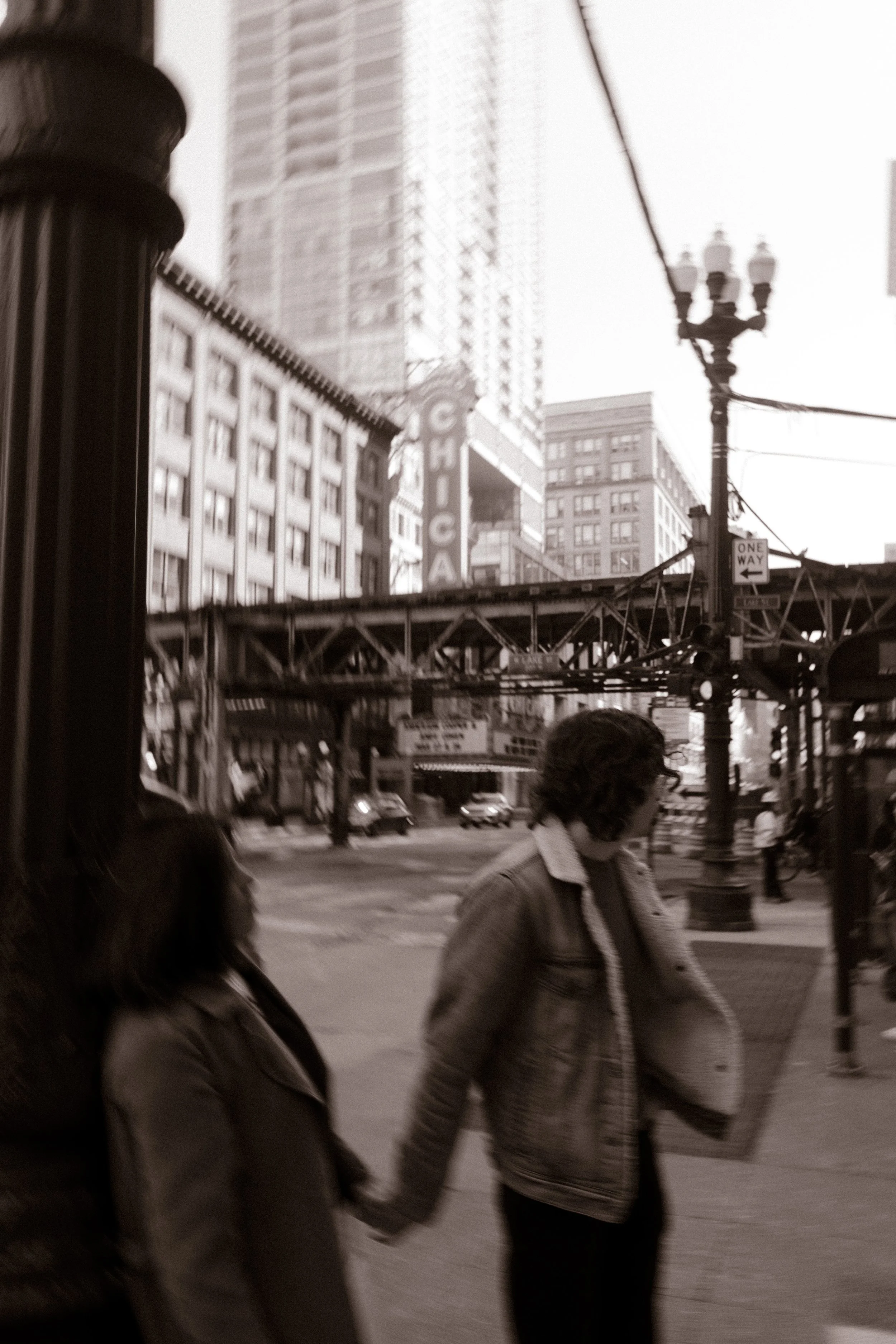 Motion blur moment of couple walking through downtown Chicago streets near the “Chicago” sign and elevated train tracks during engagement photos in Chicago
