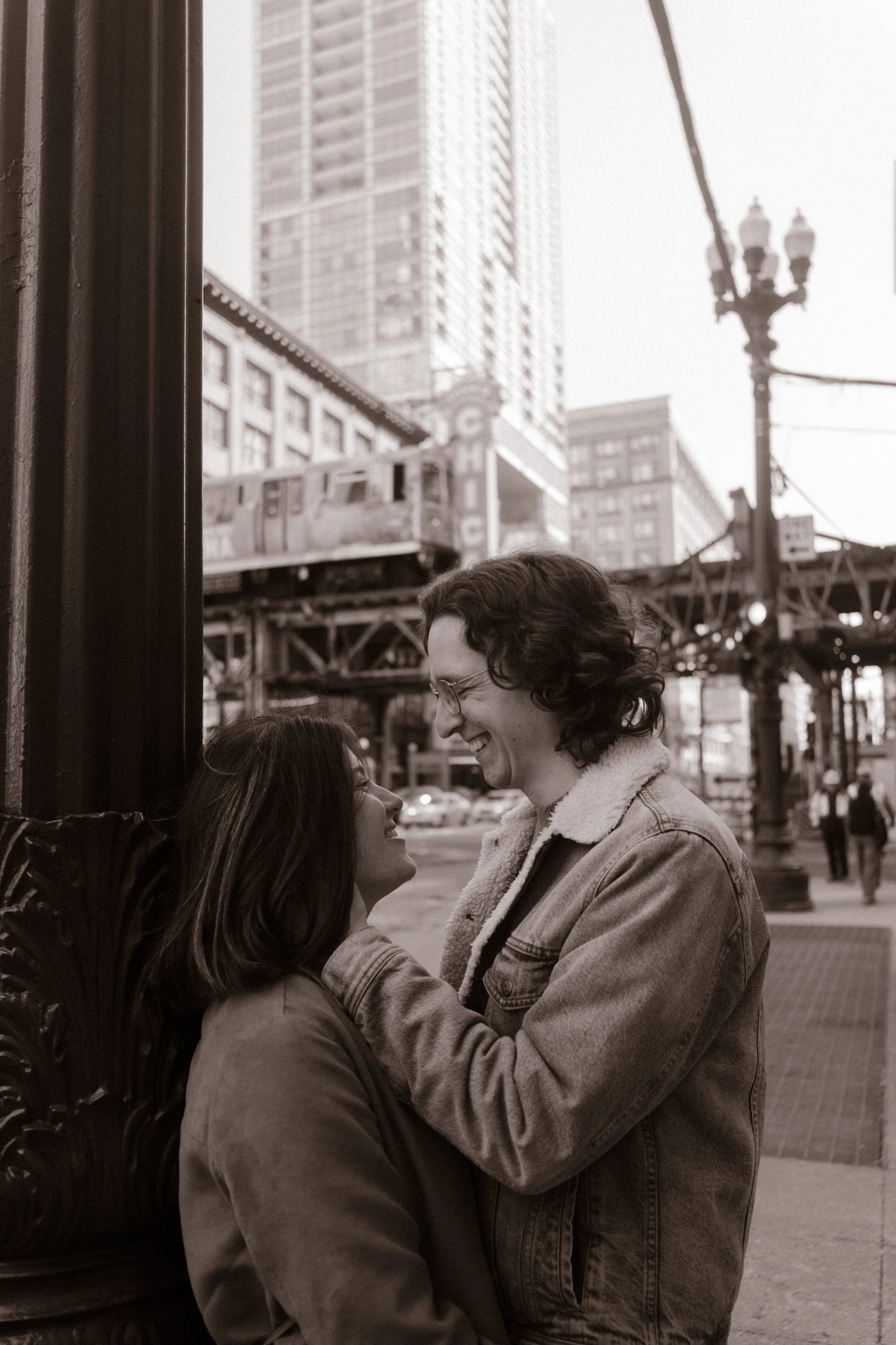 Black and white photo of couple smiling at each other near the Chicago Theatre and elevated train tracks during engagement photos in Chicago