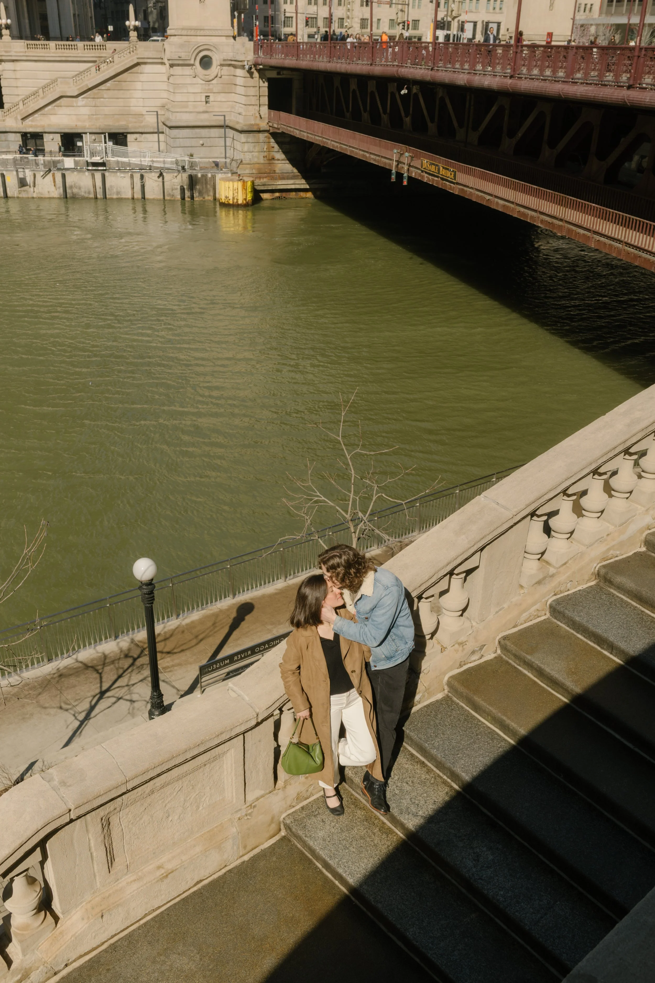 Overhead view of couple kissing on stone steps near the riverwalk, framed by bridge architecture and water below