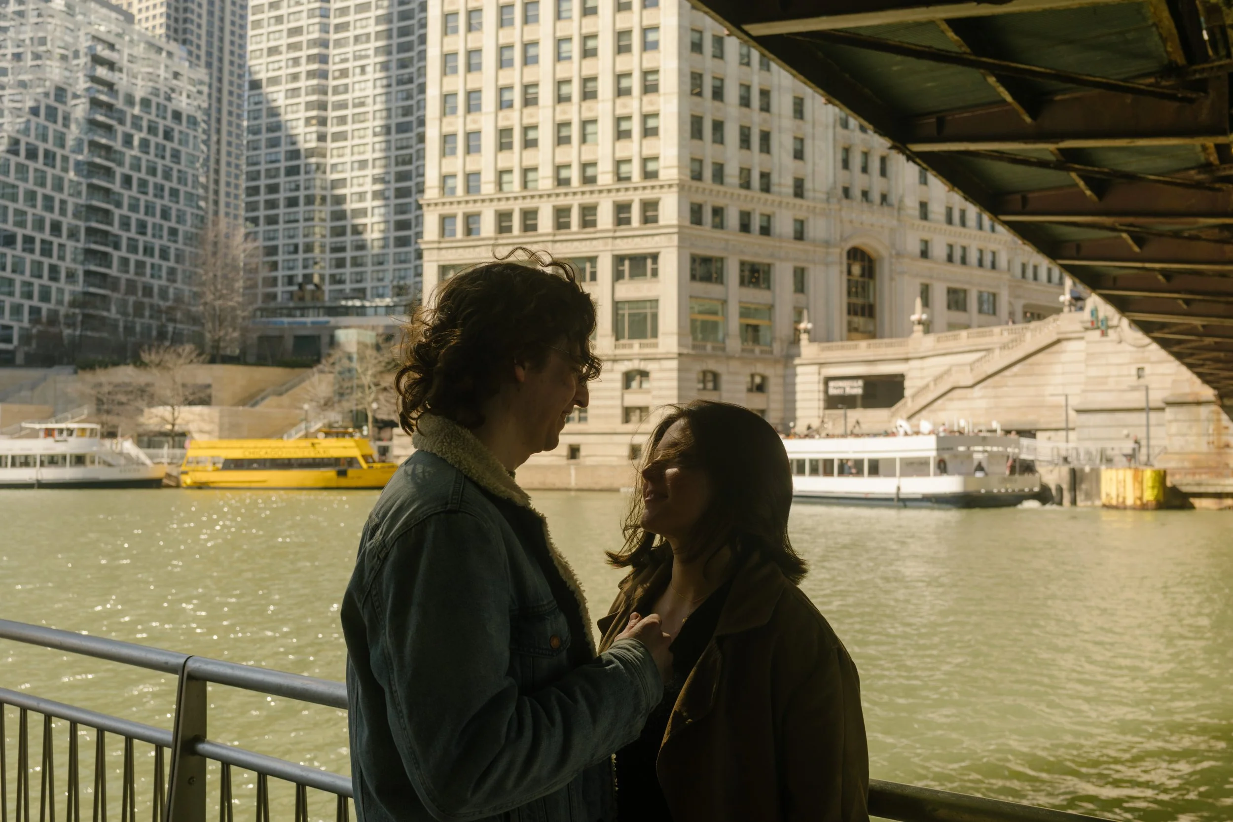 Close-up of couple laughing together under a bridge along the riverwalk, city buildings and water behind them