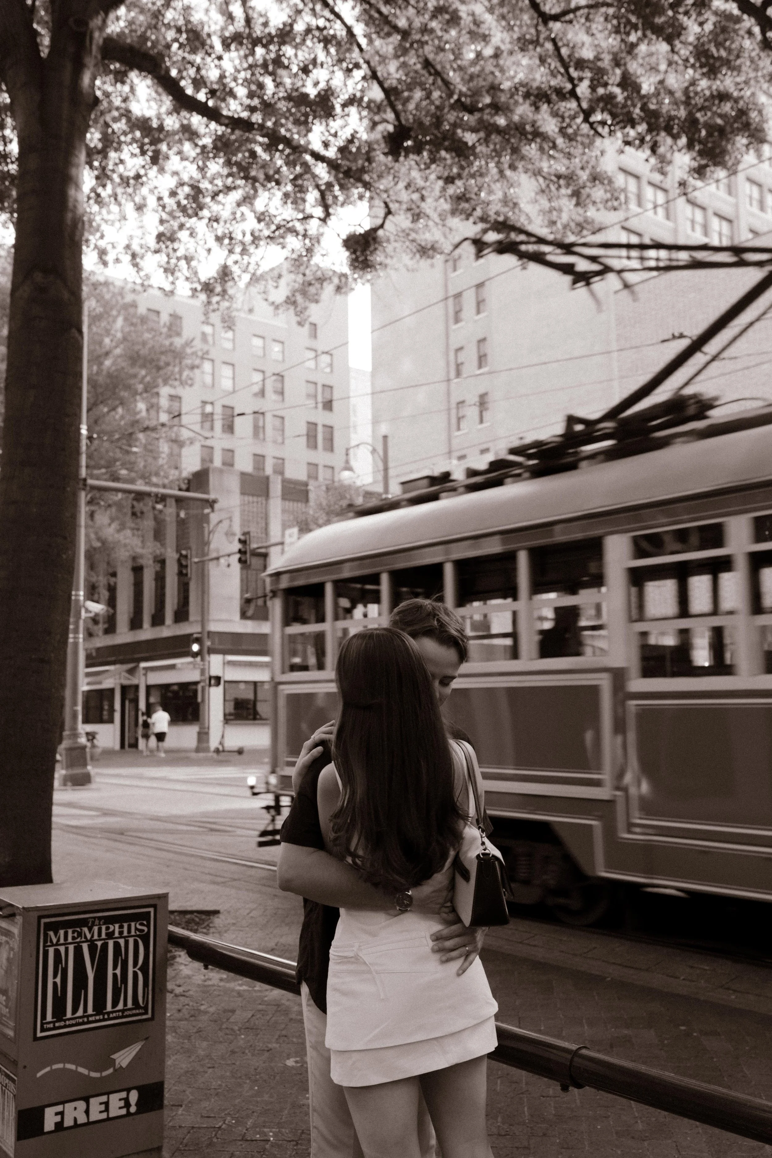 Black and white image of couple hugging near a passing streetcar in a downtown environment with a documentary feel