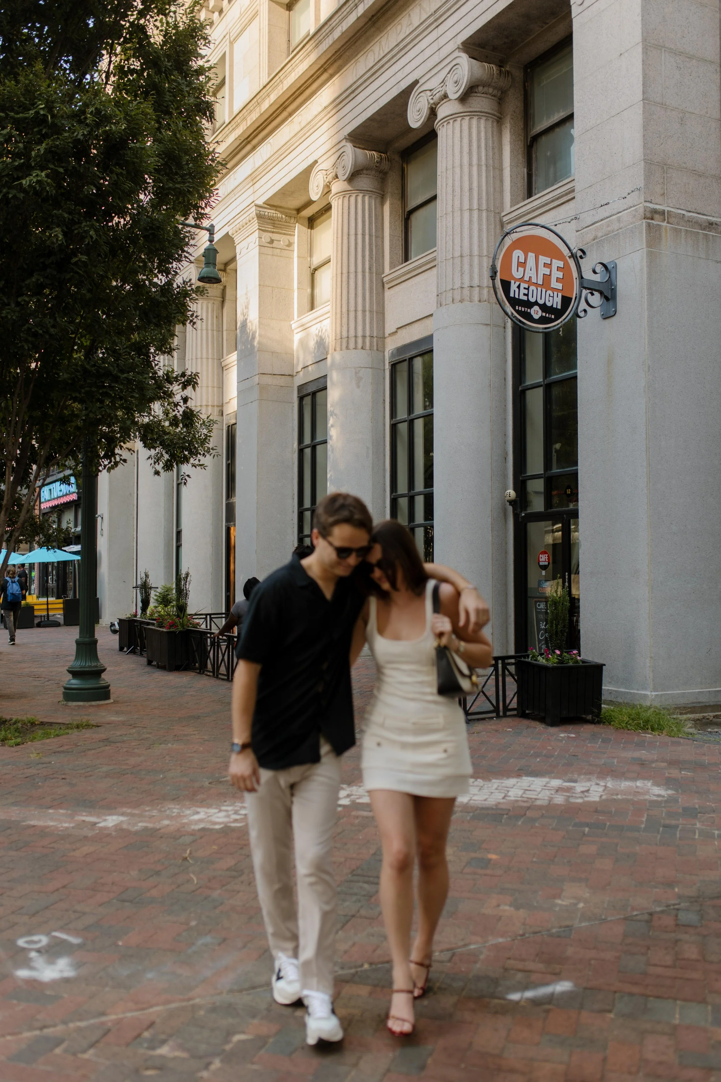 Couple walking arm in arm past a cafe on a city street during new york city engagement photos with soft afternoon light