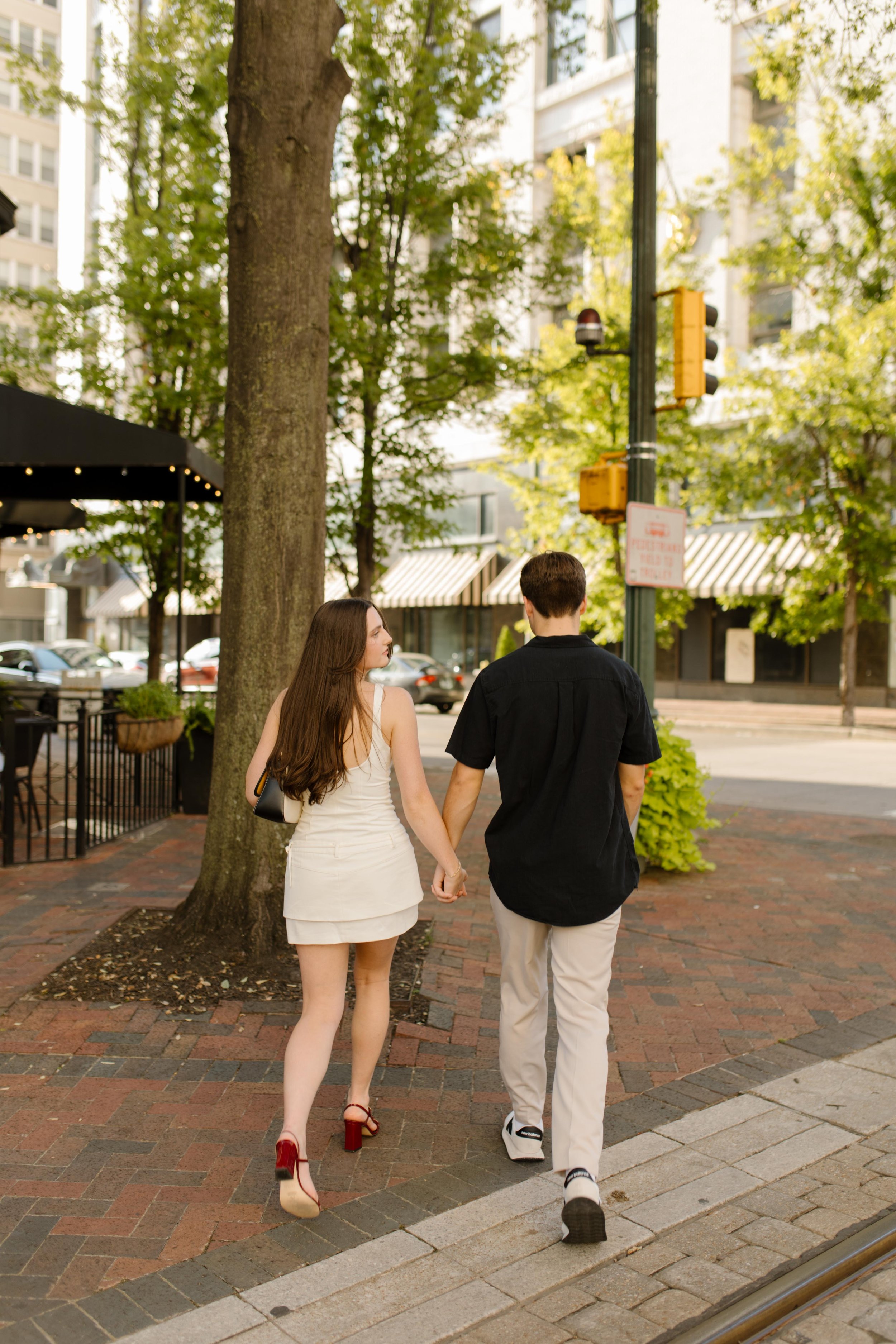 Couple walking hand in hand past trees and storefronts in a relaxed urban engagement session