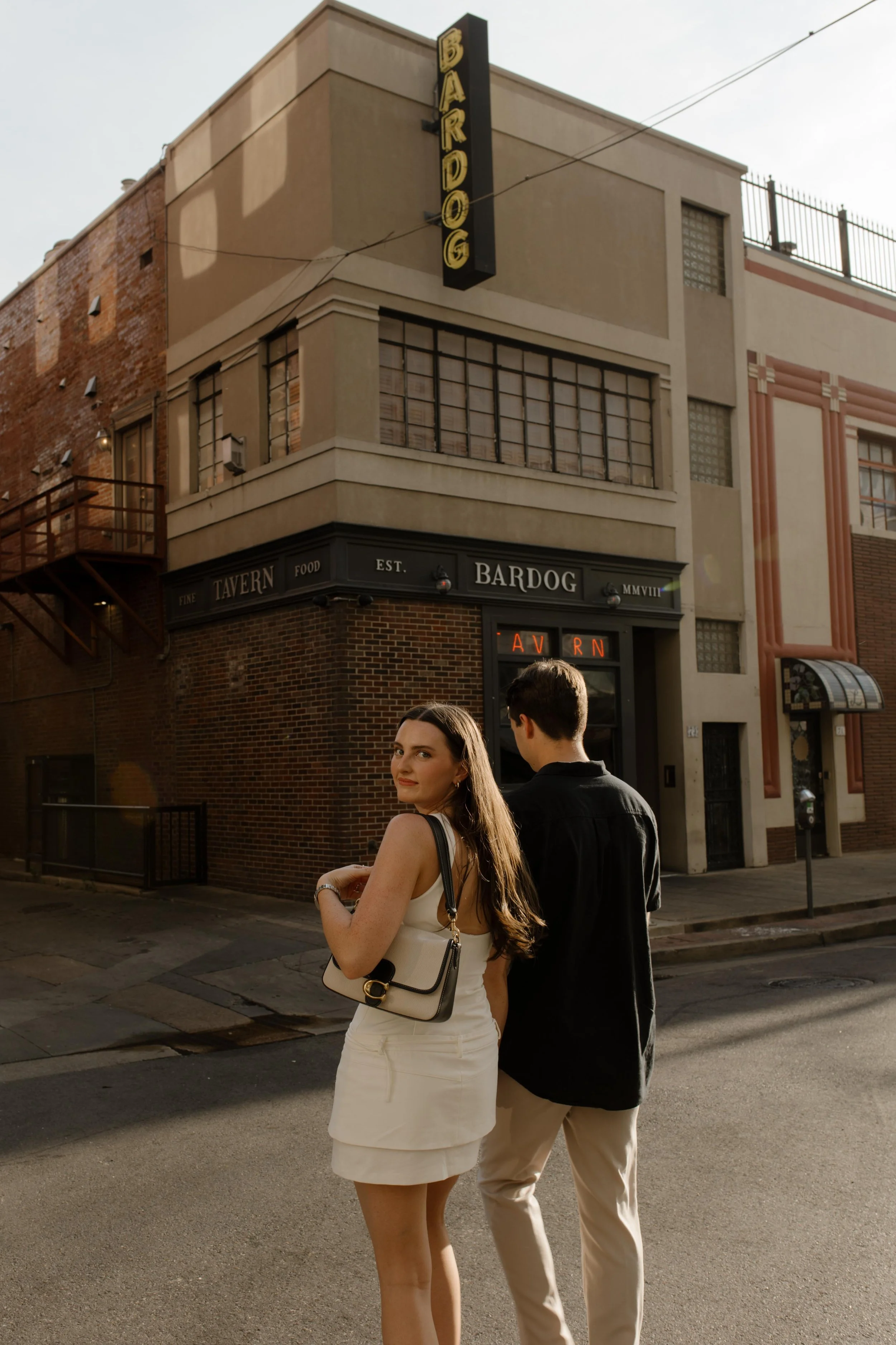 Couple standing at a street corner with warm light and a city backdrop featuring brick buildings and storefronts