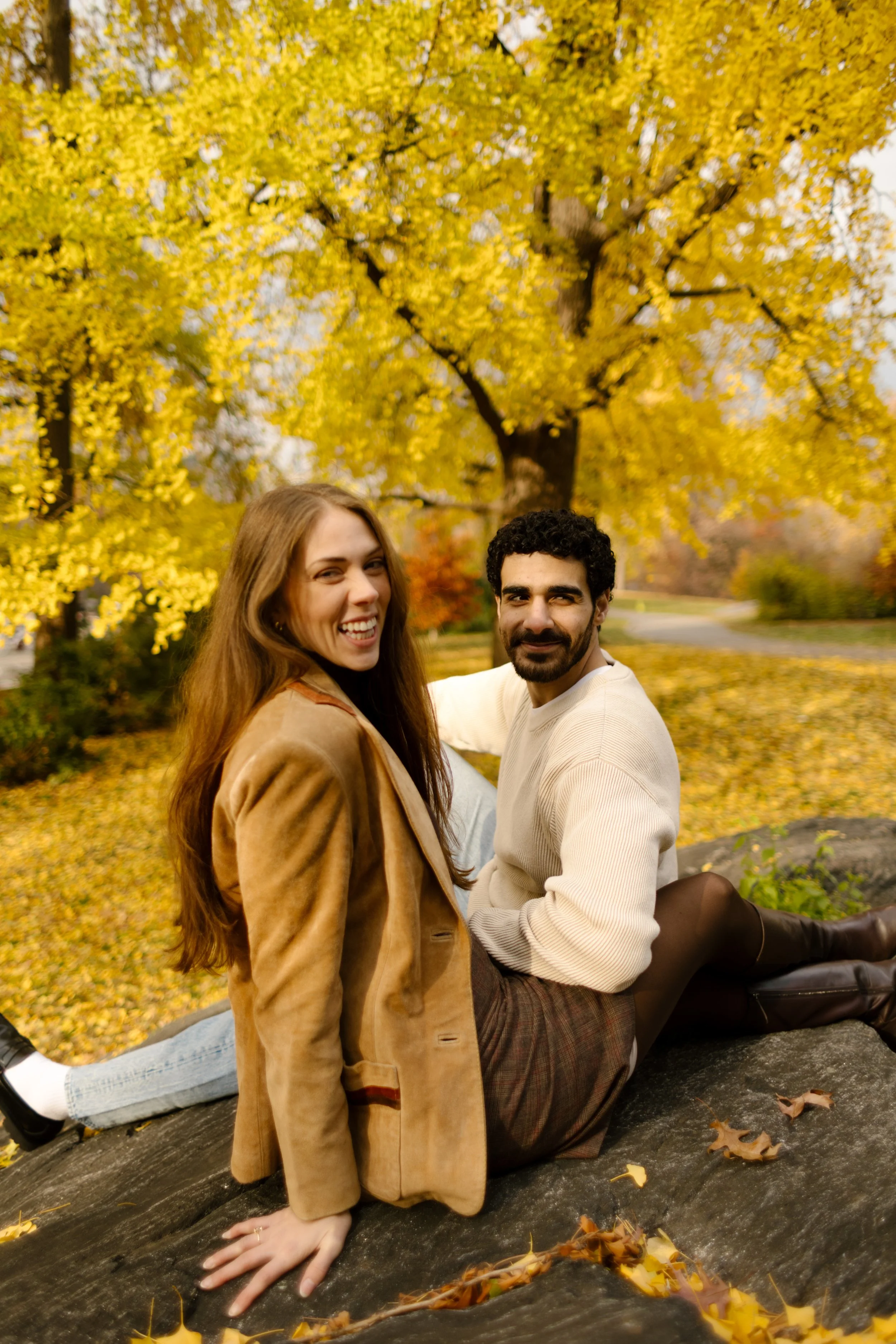 Couple sitting together on a rock surrounded by vibrant fall leaves in Central Park during an engagement session.