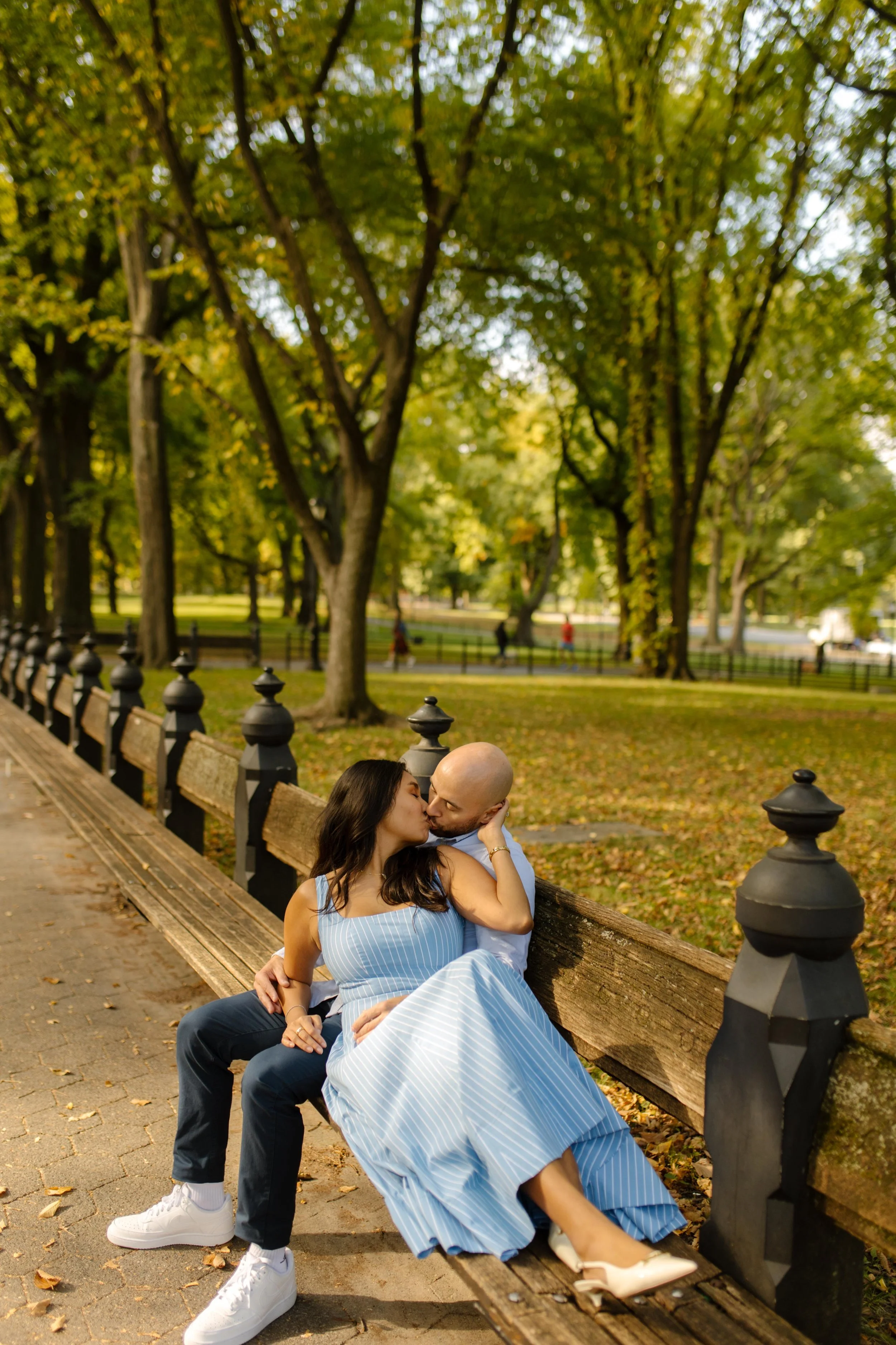 Couple kissing on a Central Park bench surrounded by trees during a relaxed autumn engagement session.