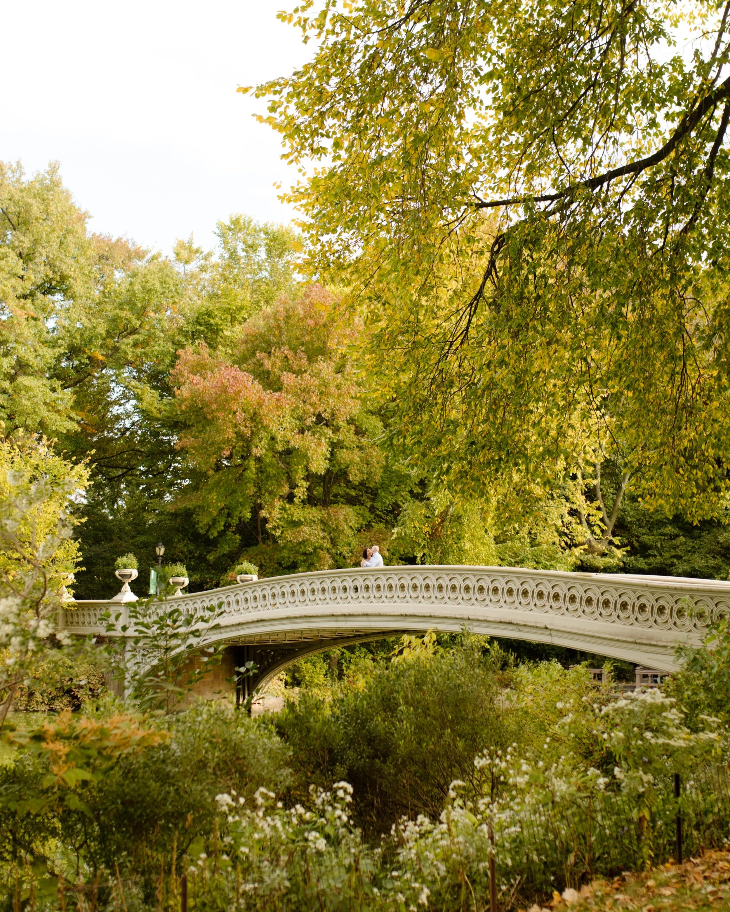 New York engagement photographer capturing a couple walking across Bow Bridge surrounded by autumn foliage in Central Park.