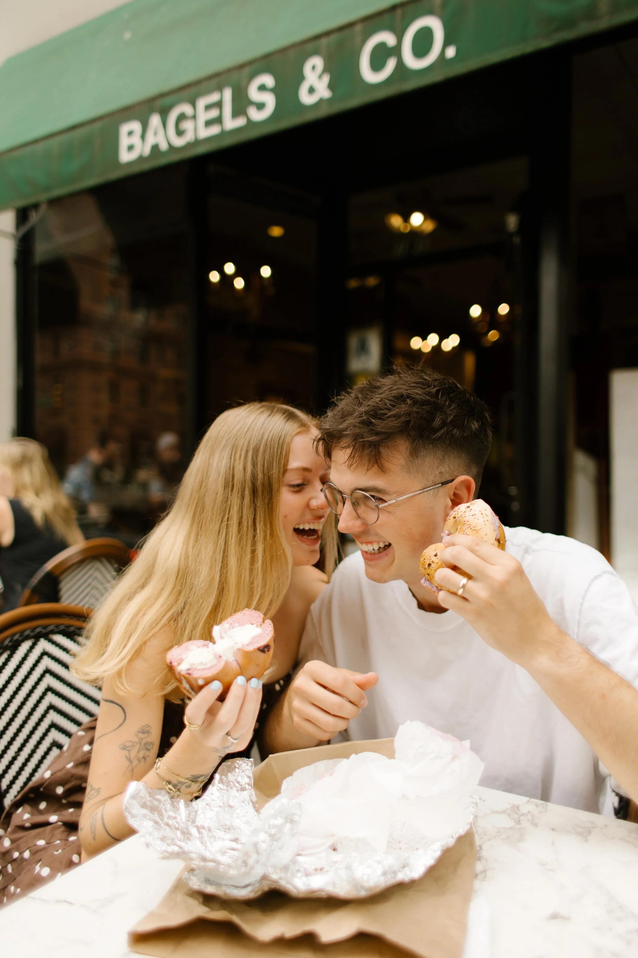 Couple laughing together over bagels and coffee outside a New York City café during their engagement session.