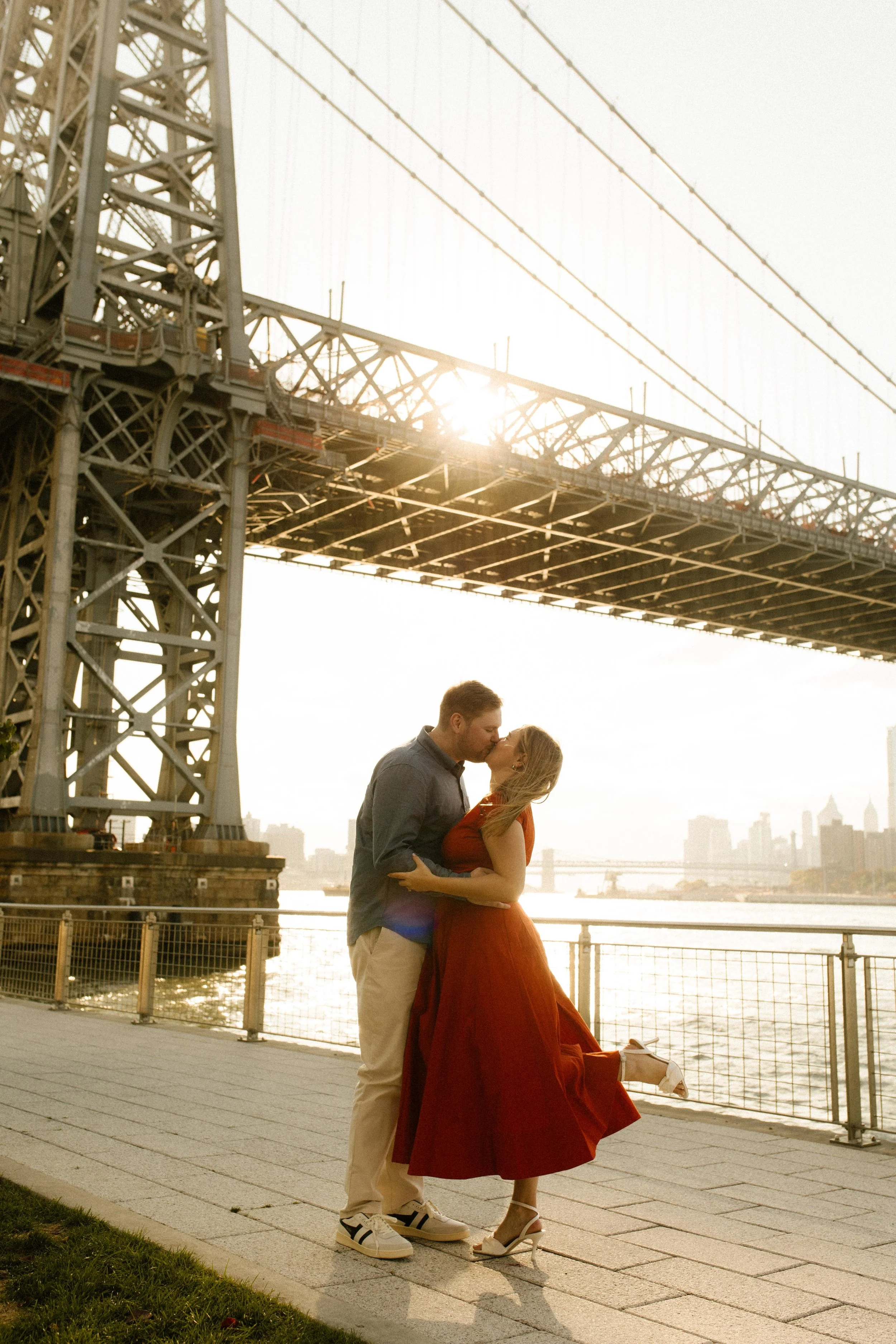 New York engagement photographer capturing a romantic waterfront kiss beneath the Williamsburg Bridge at sunset.