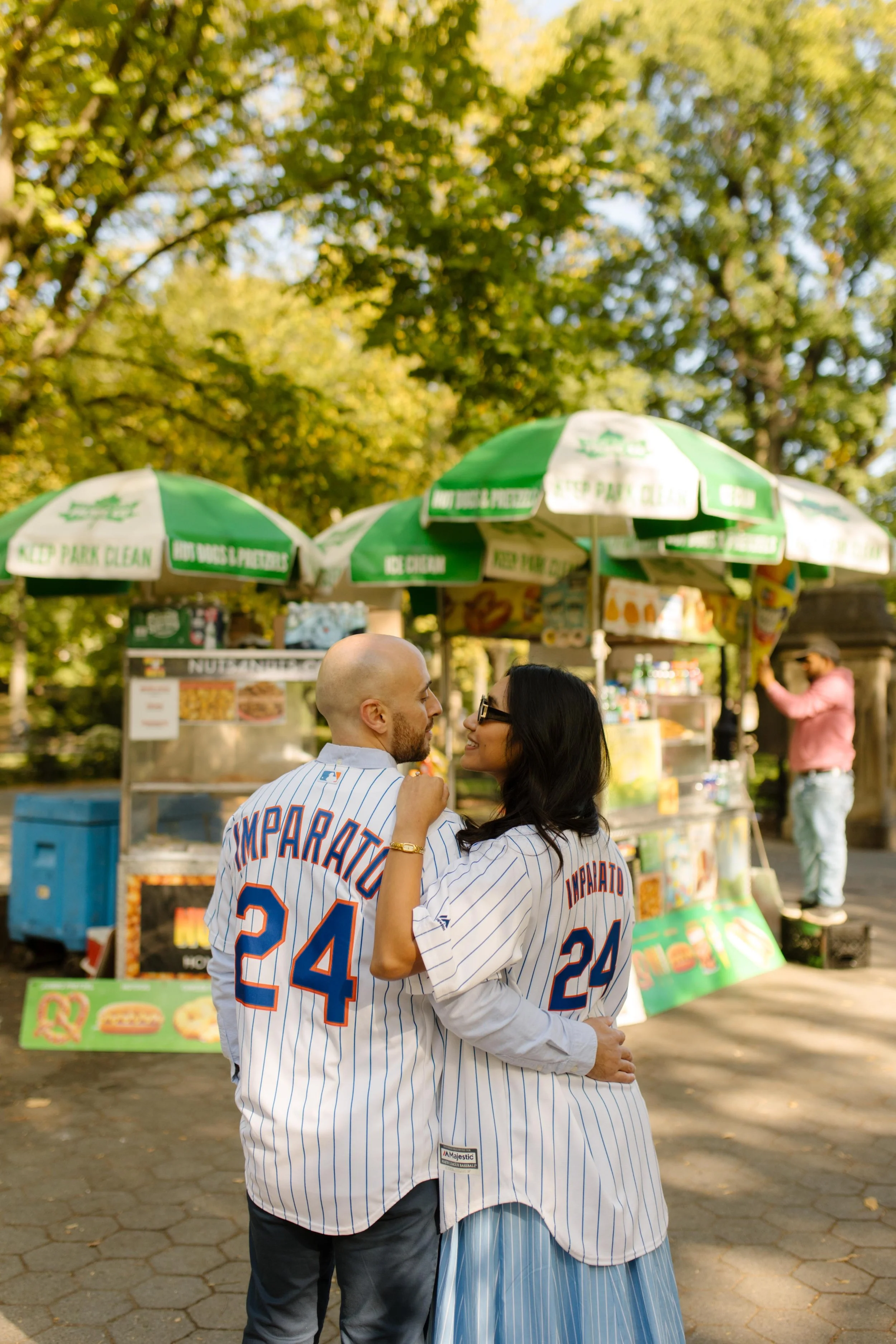 Couple embracing in matching Mets jerseys near a Central Park food stand during a casual engagement session.