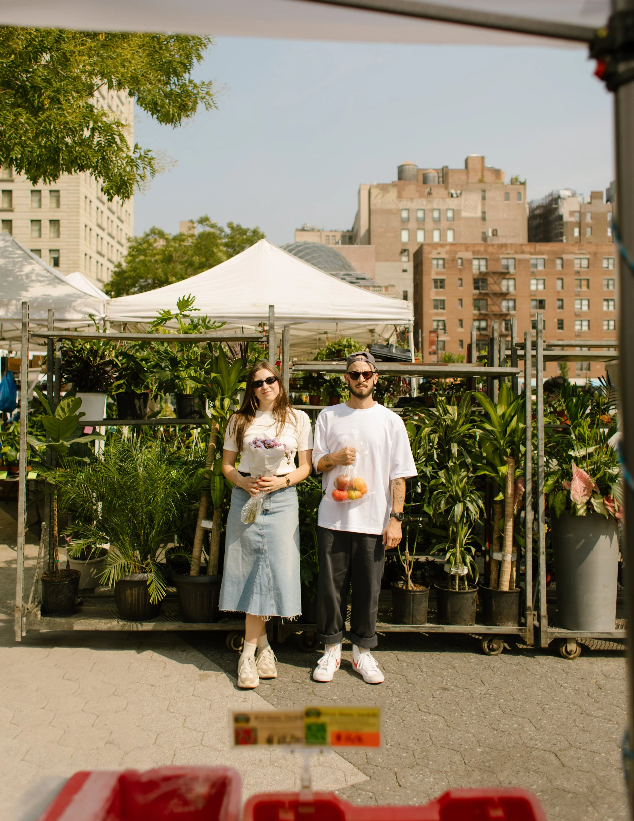 Couple posing with flowers and fresh produce at a Manhattan farmers market during a relaxed city engagement session.