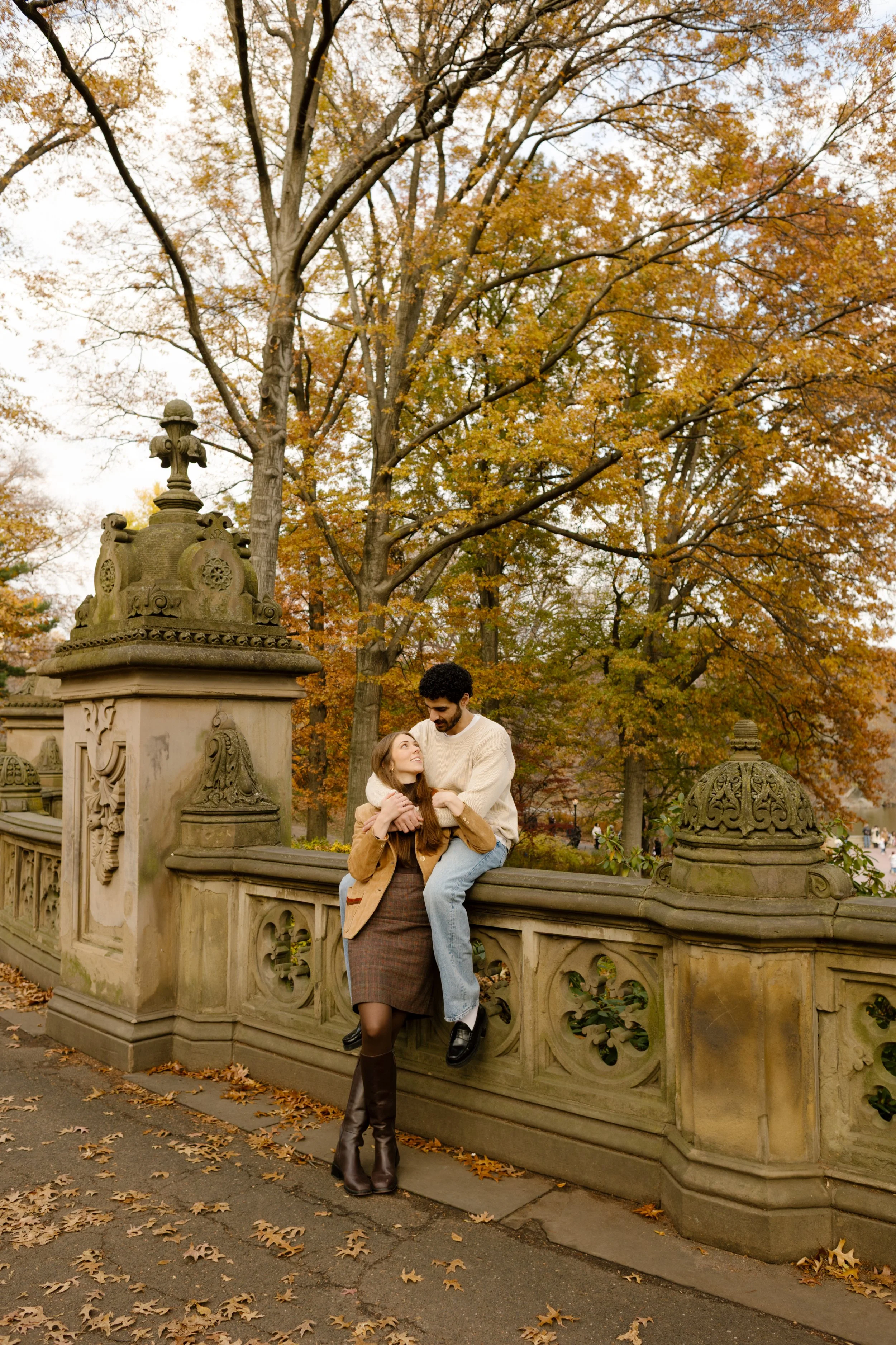New York engagement photographer capturing a couple cuddling on the Bow Bridge in Central Park surrounded by fall foliage.