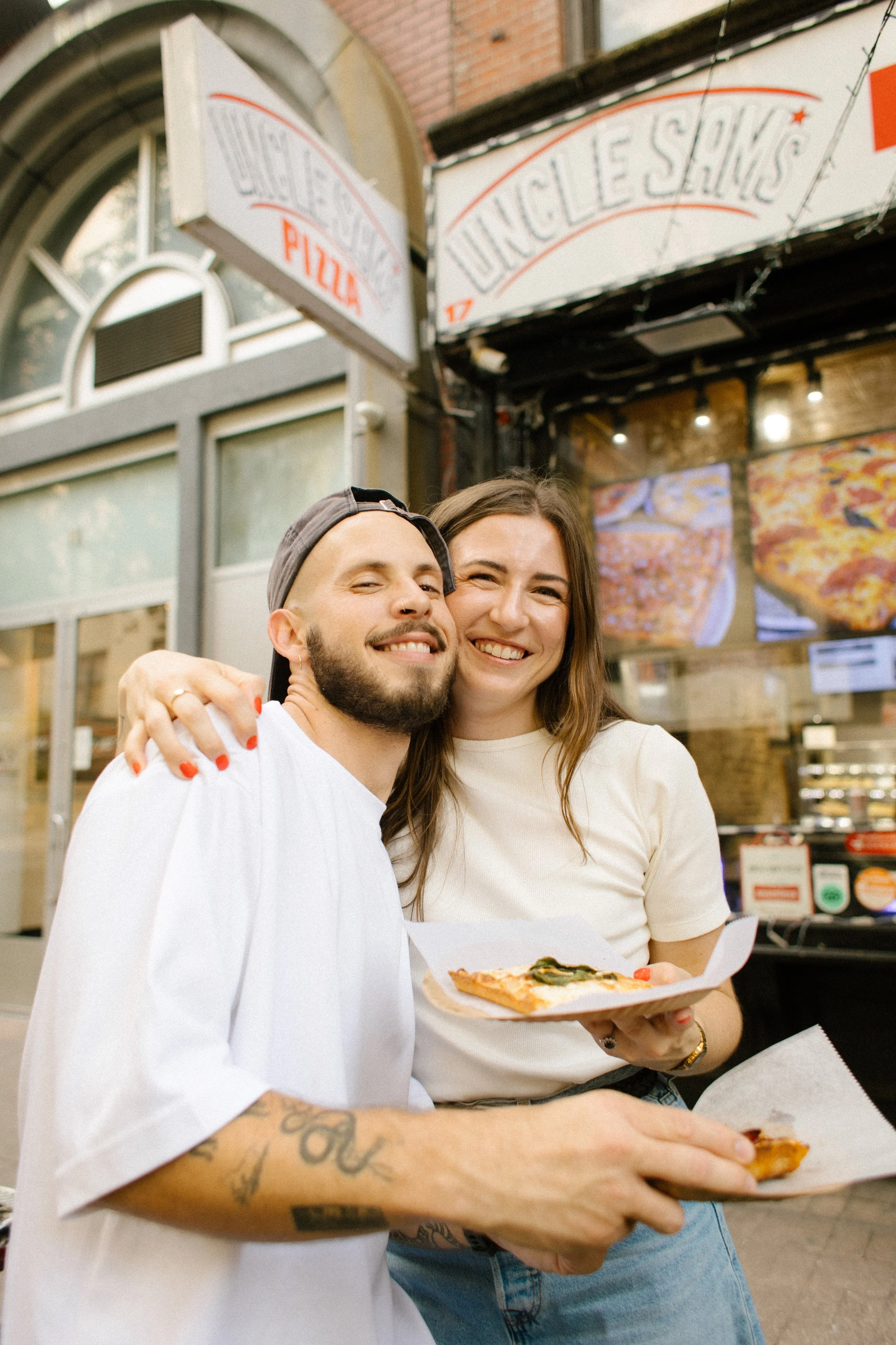 New York engagement photographer documenting a couple smiling together outside a neighborhood pizza shop in New York City.