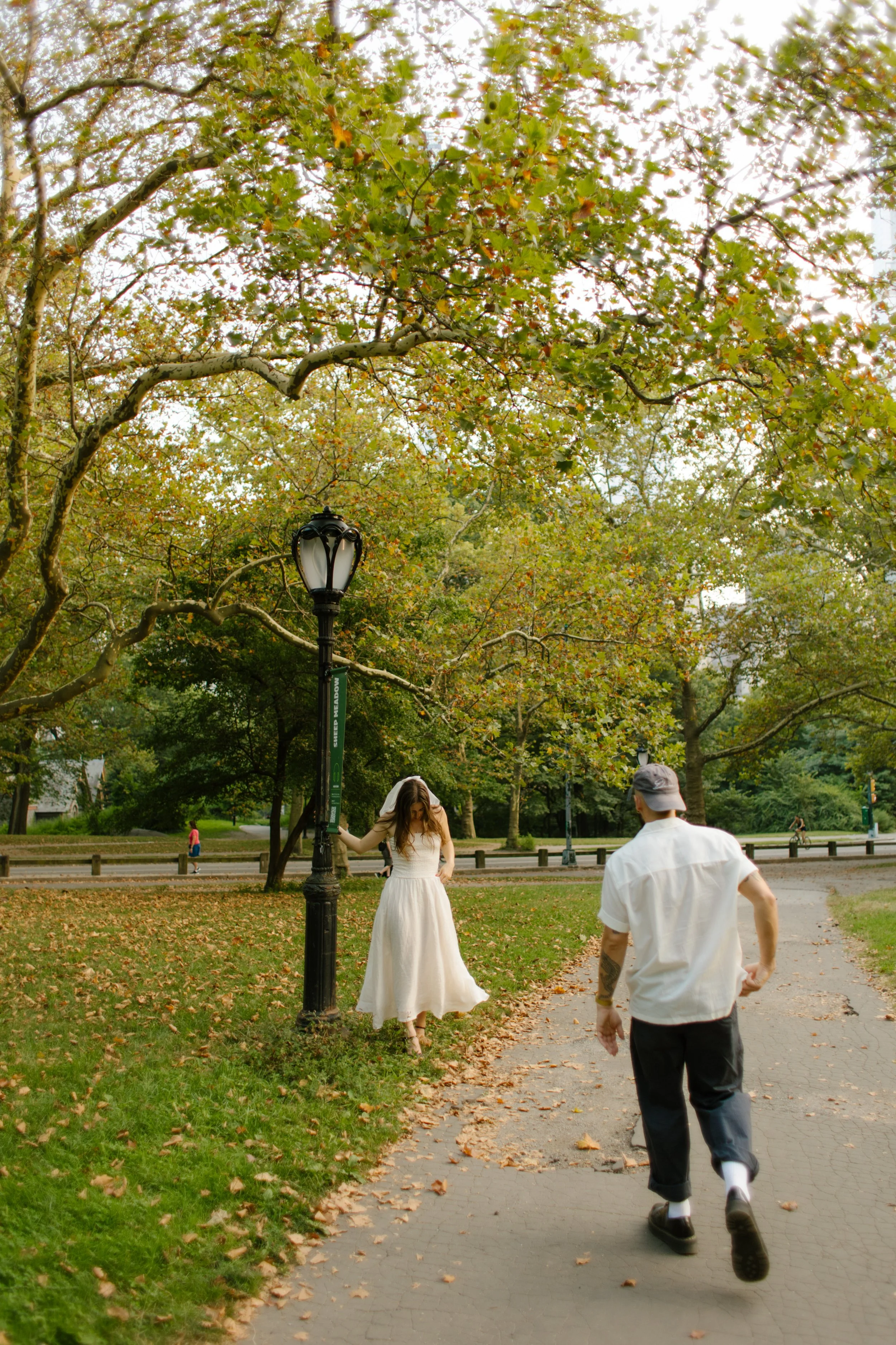 Couple running through Central Park beneath green autumn trees during a playful engagement session.