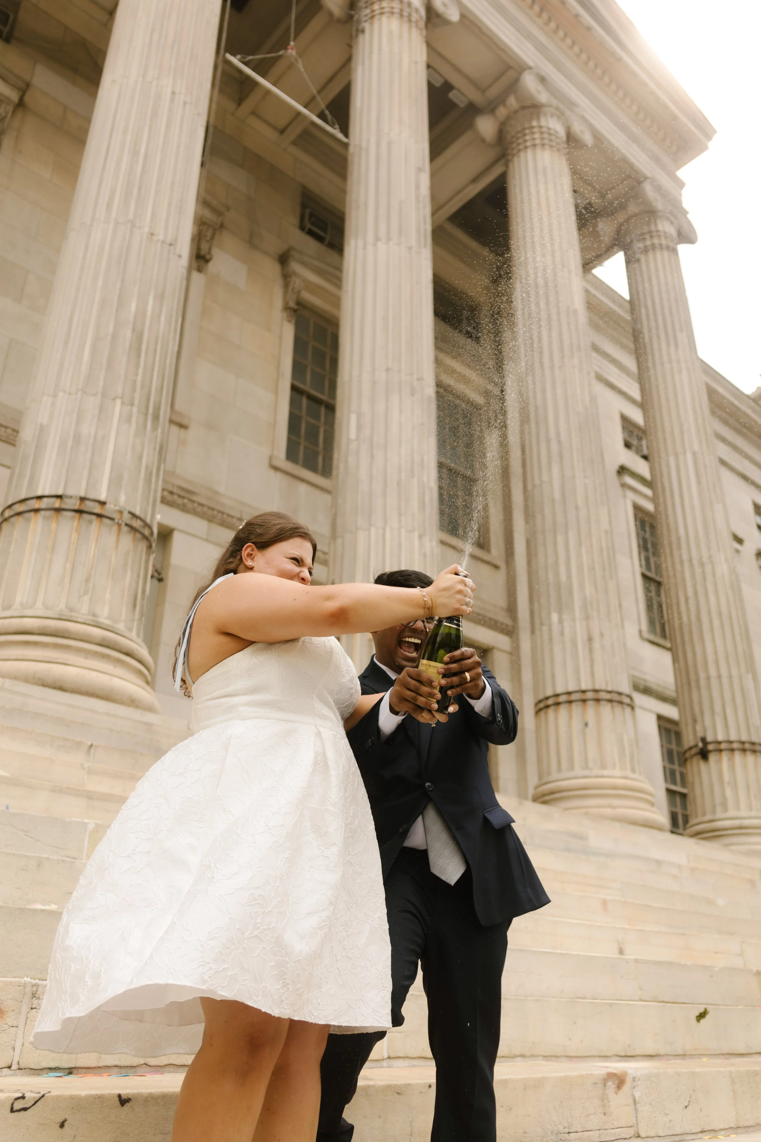 Newlyweds opening champagne outside grand courthouse steps after intimate wedding ceremony