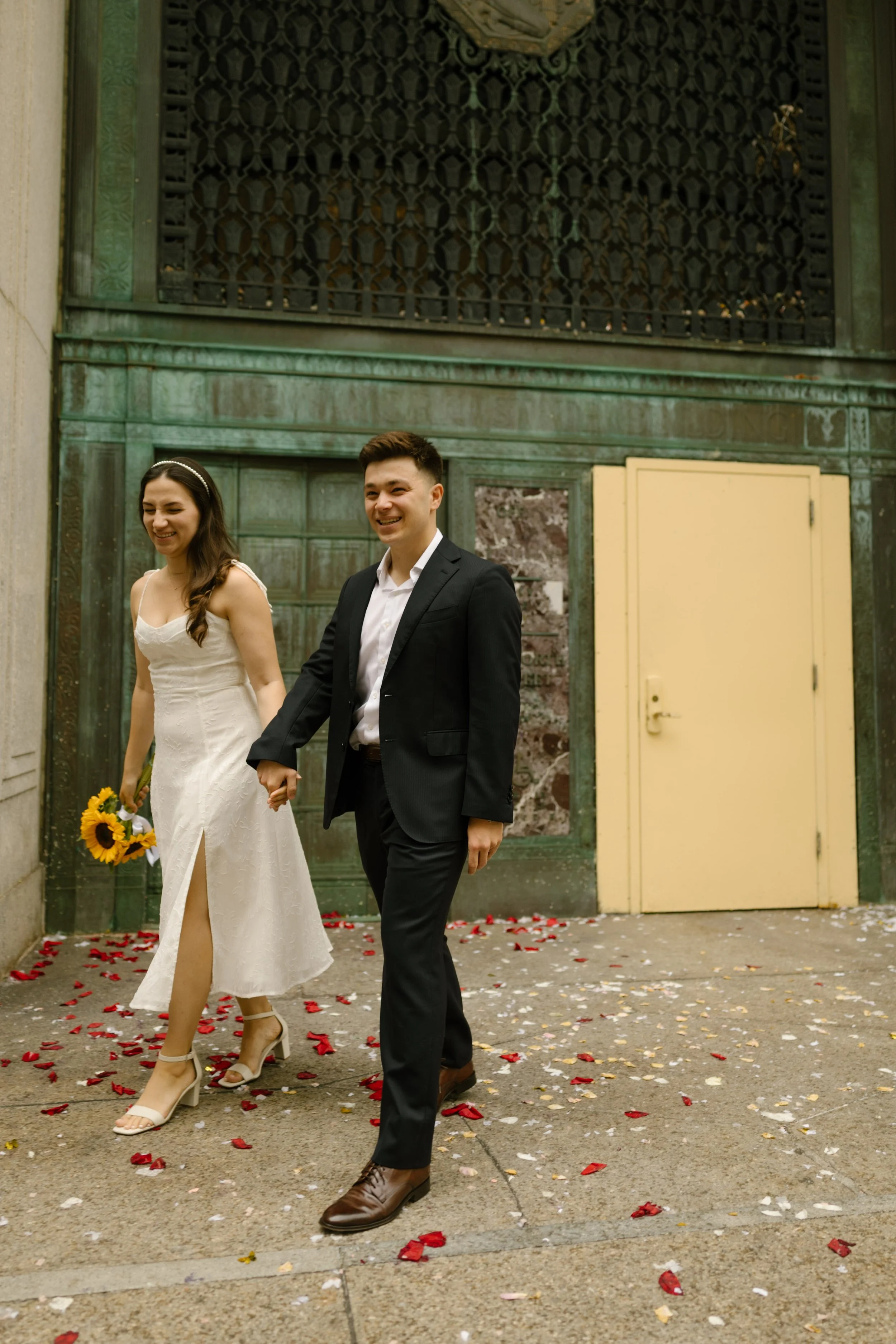 Newlyweds holding hands and walking through flower petals outside Manhattan courthouse