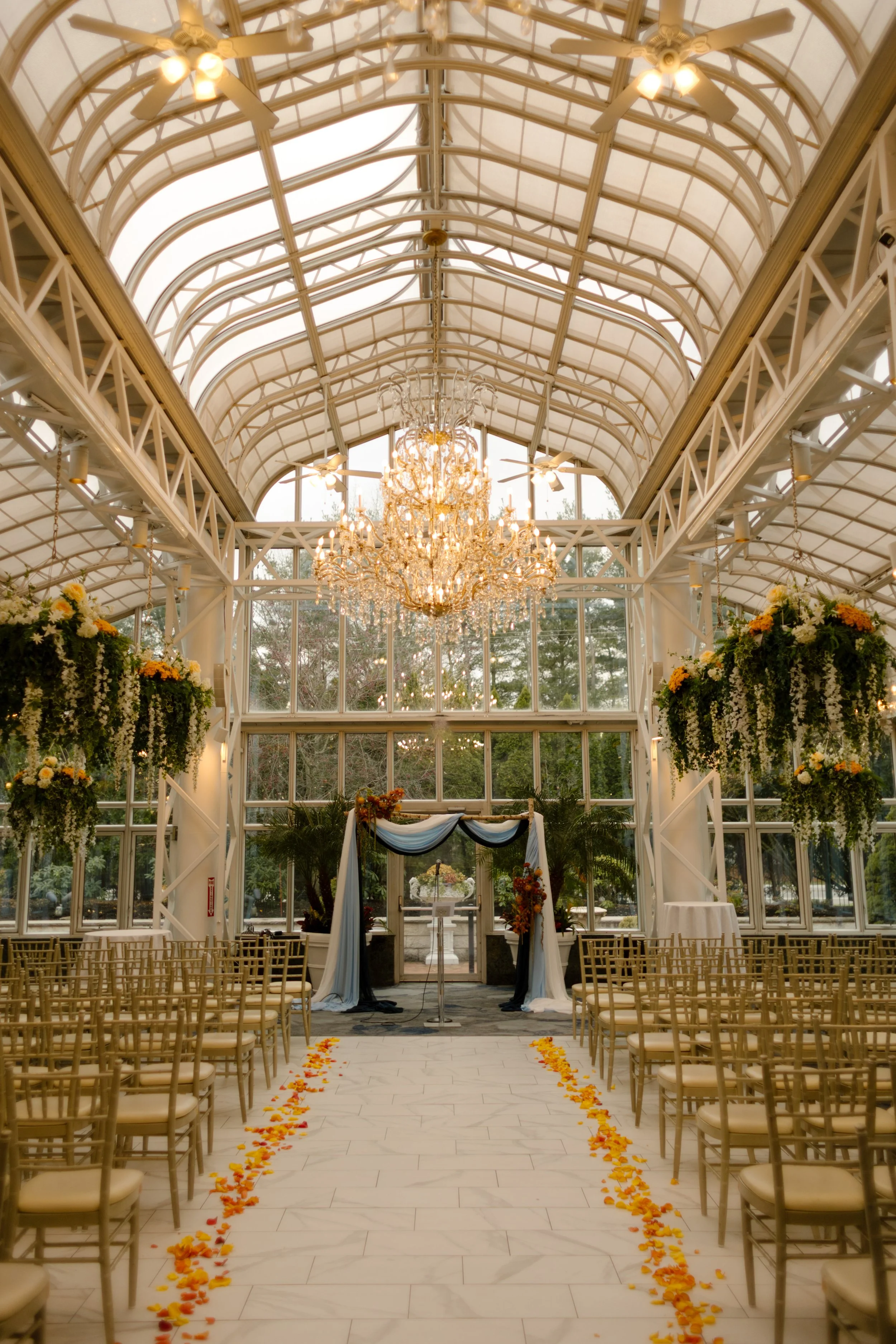 Elegant glass conservatory ceremony space with chandeliers, floral installations, and rows of guest seating