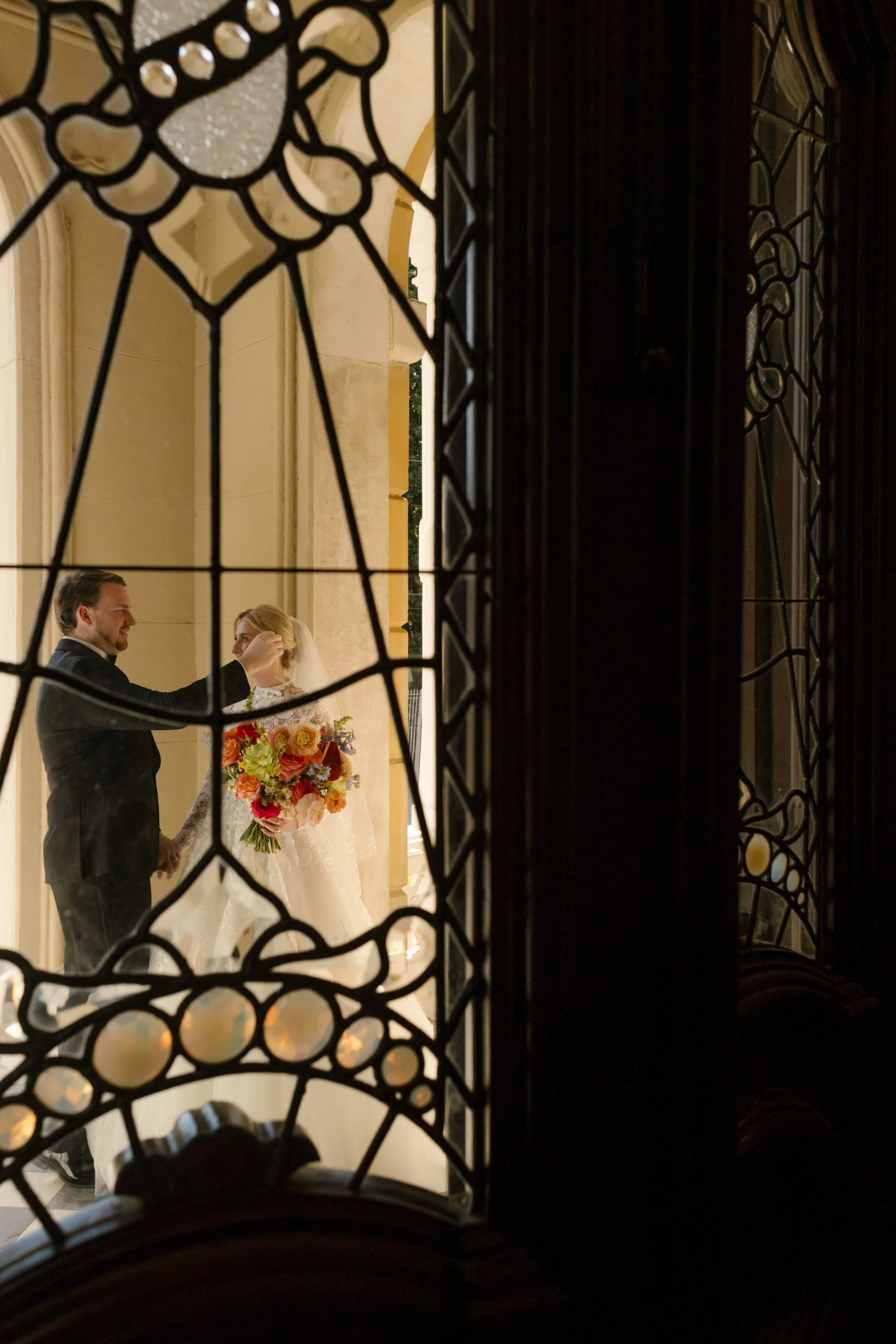 Bride and groom framed through stained glass with vibrant wedding bouquet