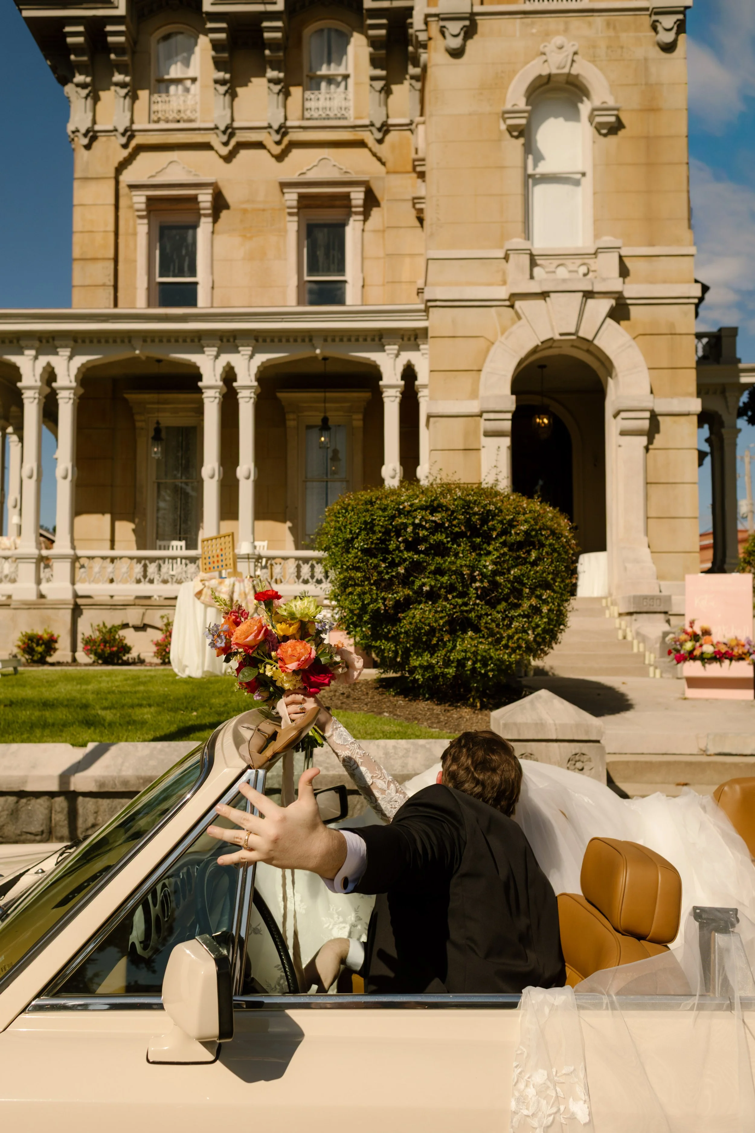 Newlyweds celebrating in a vintage convertible outside a grand historic mansion