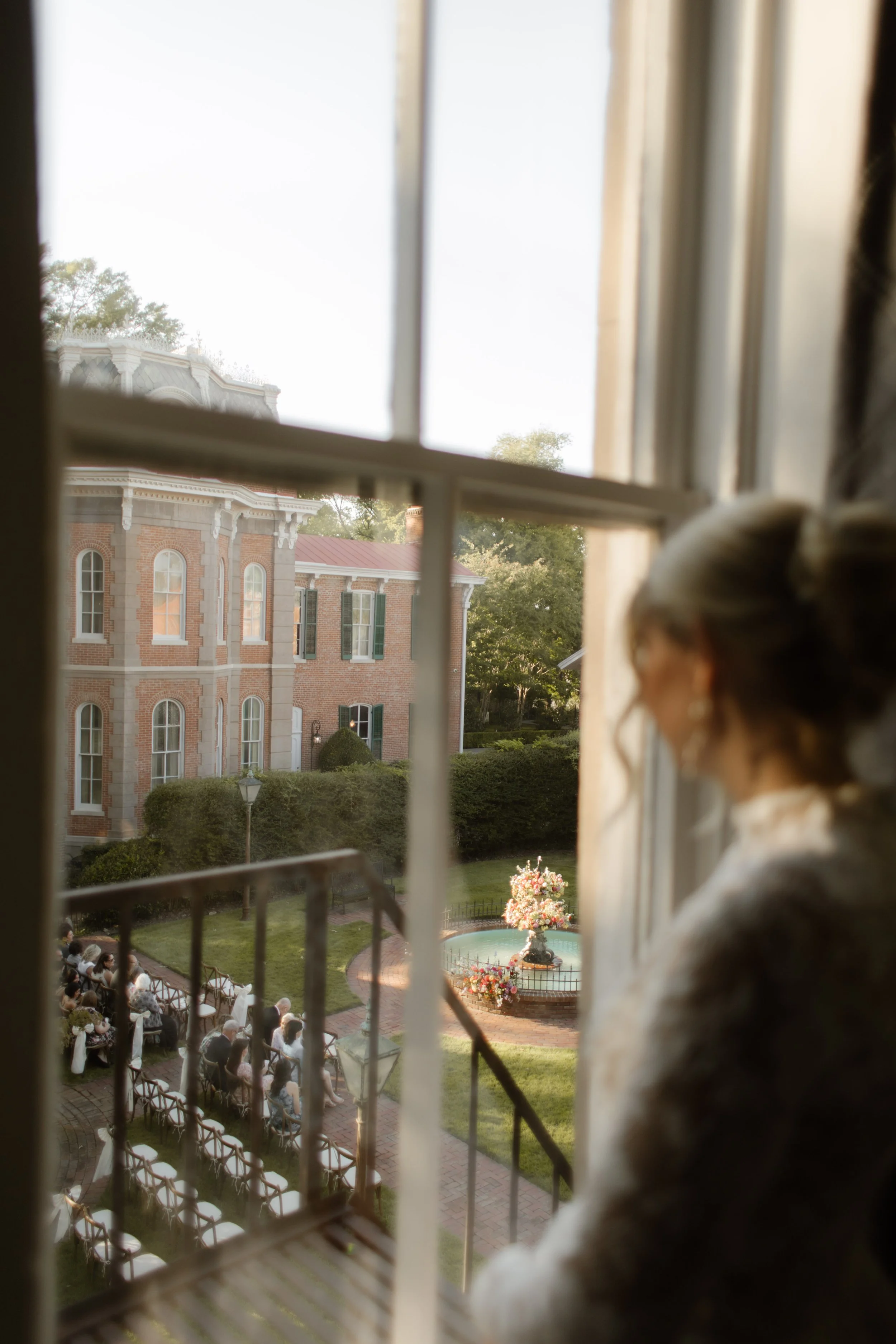 Bride watching her outdoor ceremony from an upstairs window before walking down the aisle