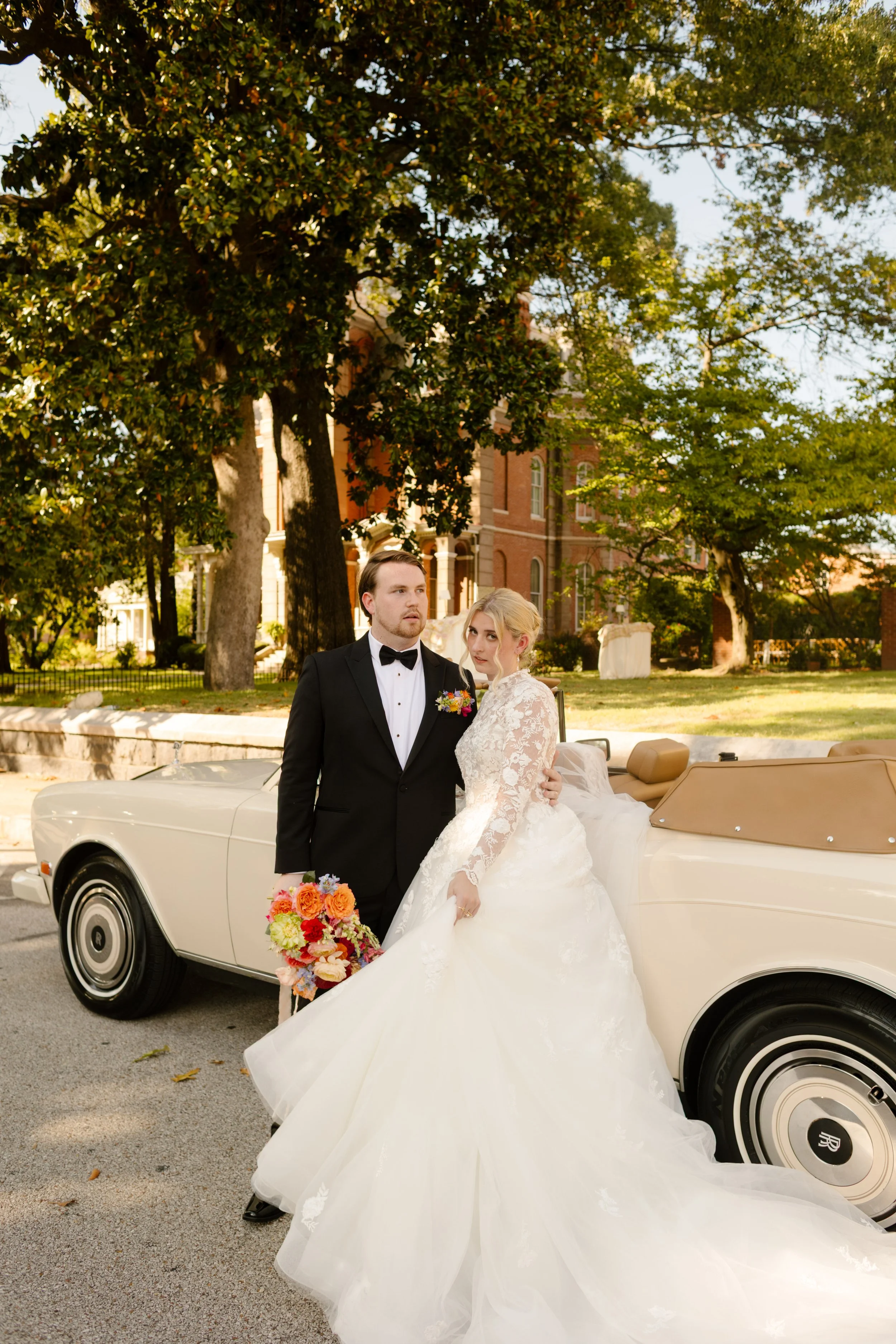 Bride and groom posing beside a vintage convertible with colorful florals outside a historic mansion