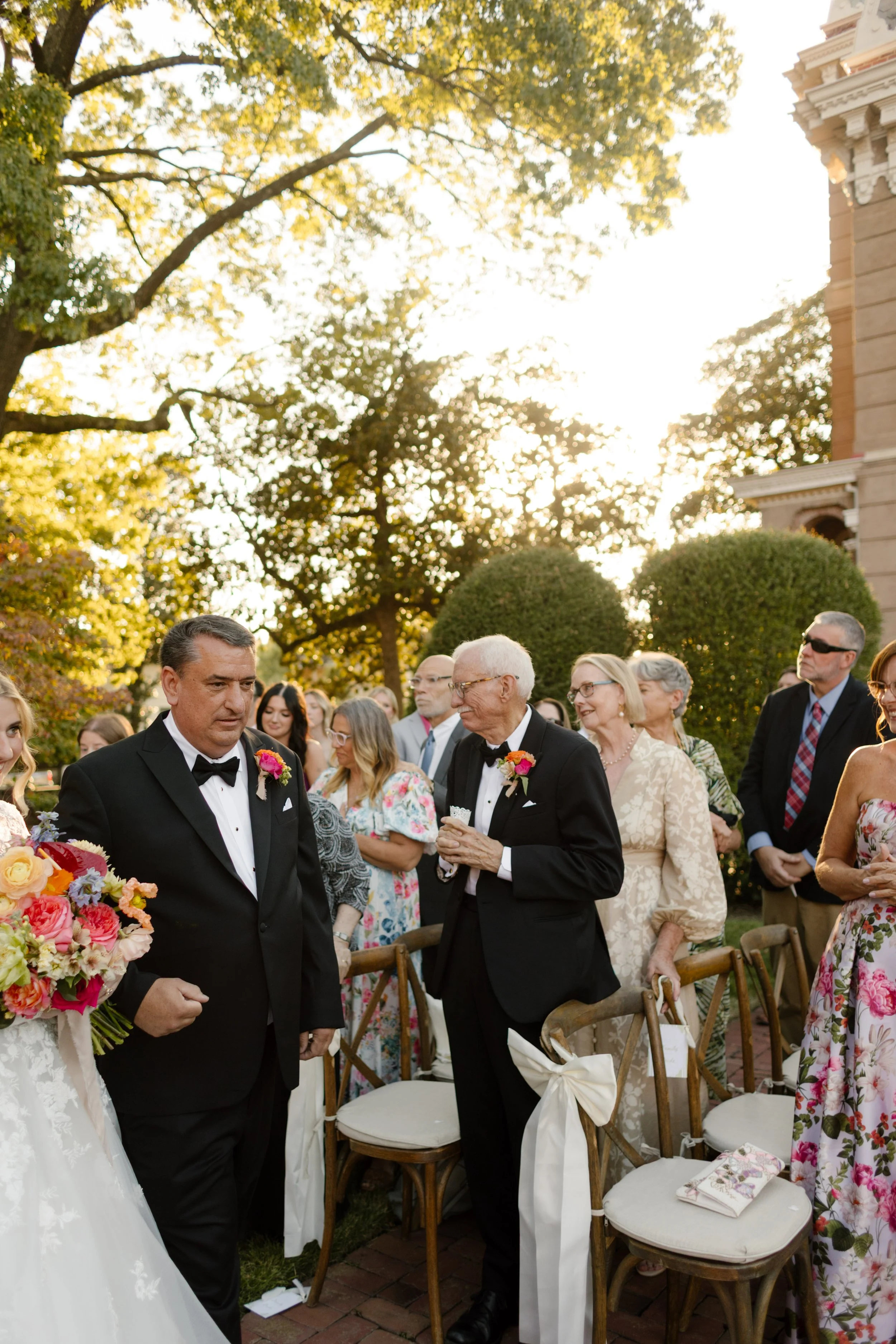 Wedding processional beginning as guests stand among garden ceremony seating under golden-hour light