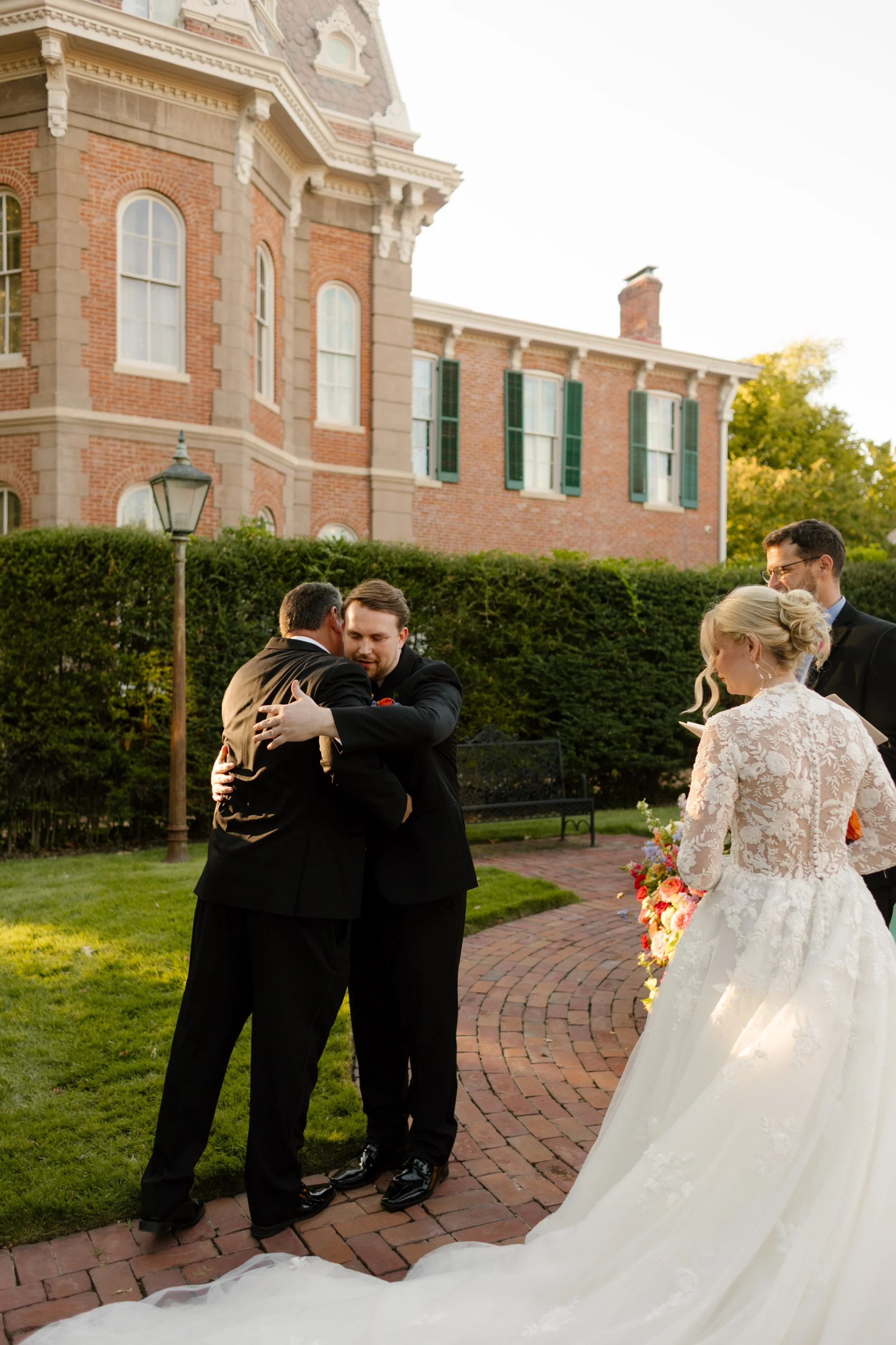 Groom embracing a family member before the ceremony while the bride waits nearby with her bouquet