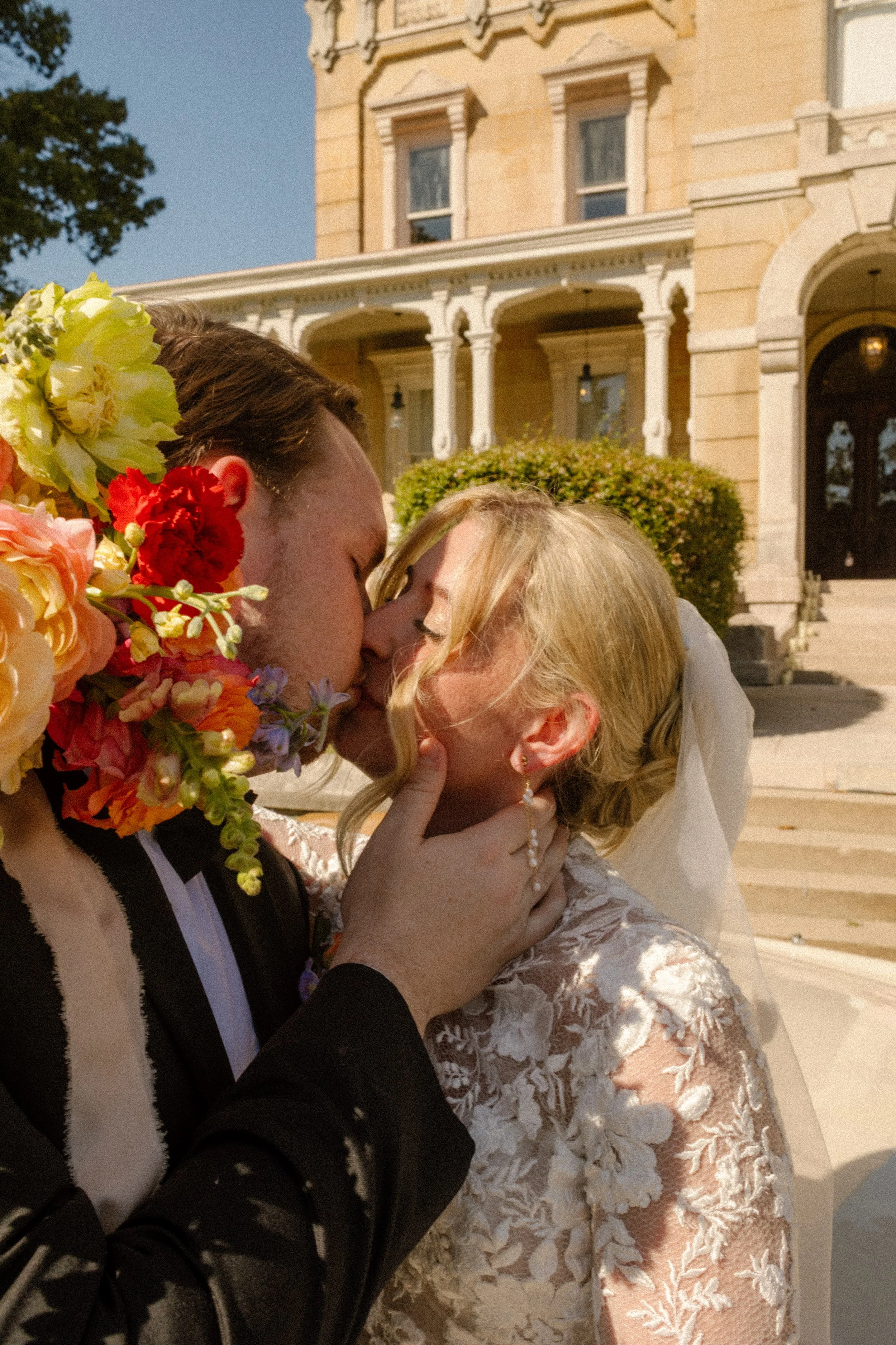 Bride and groom sharing a kiss in front of a grand mansion with lush garden-inspired wedding flowers
