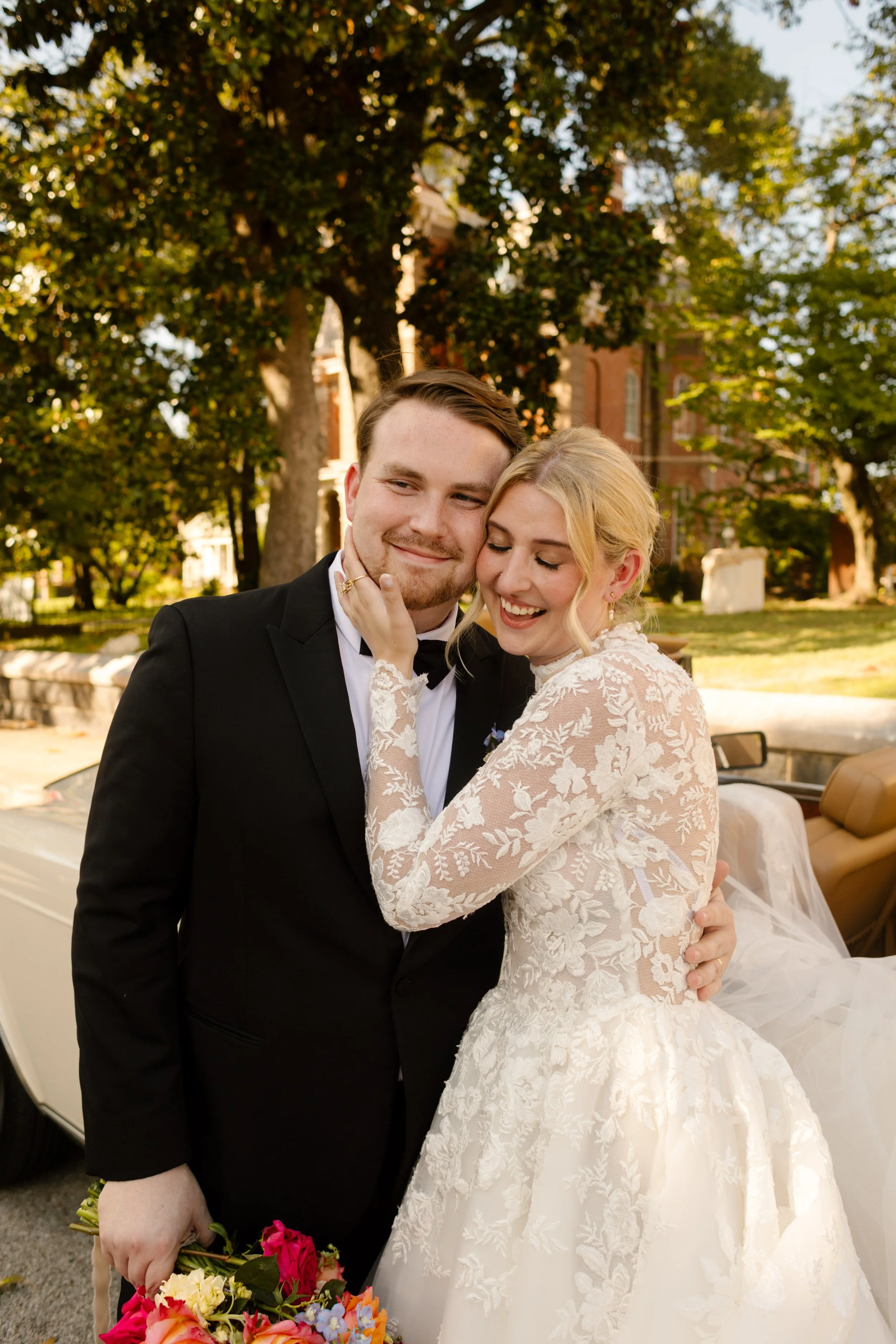 Bride and groom posing beside a classic convertible with a romantic garden backdrop and vibrant wedding florals