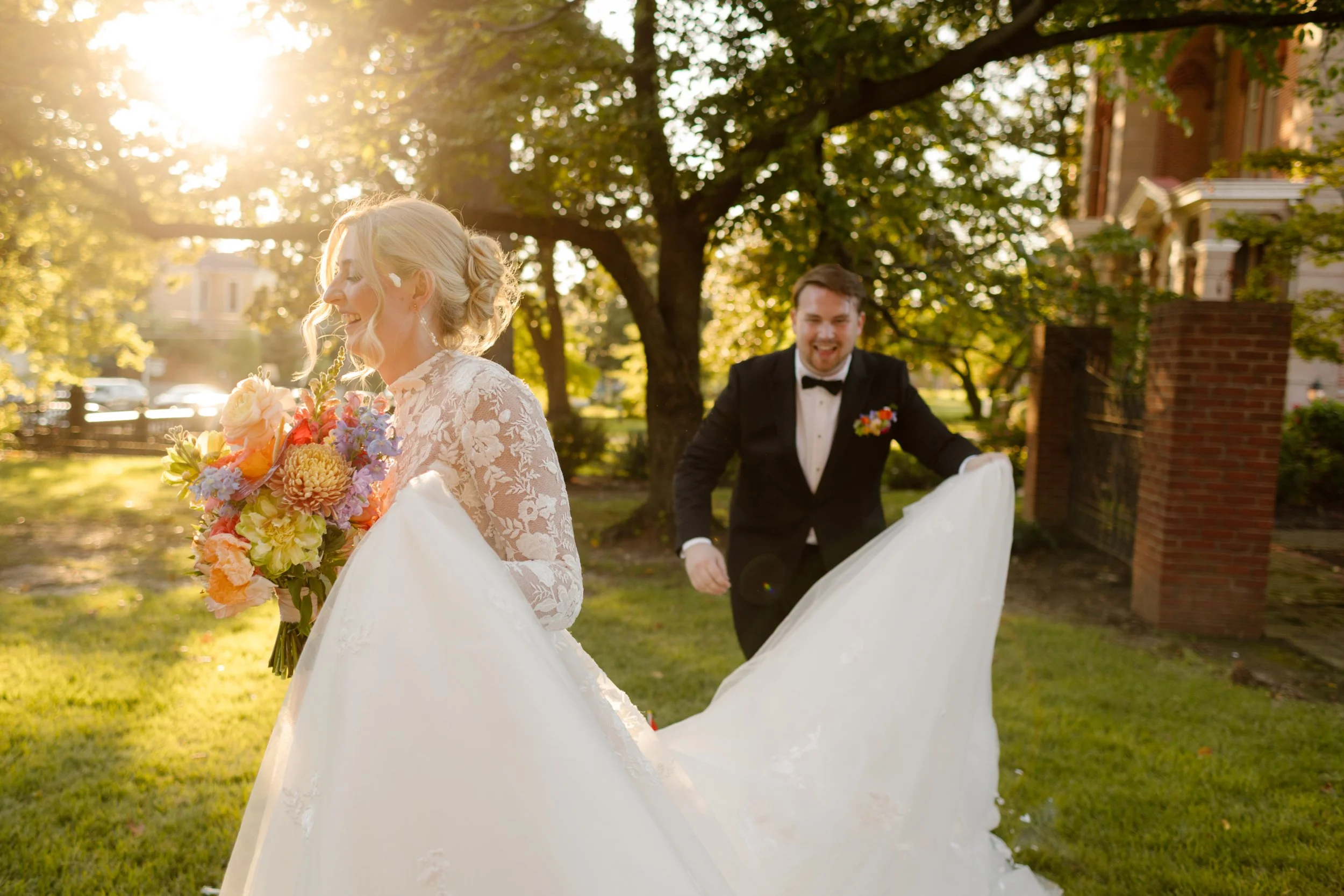 Groom playfully lifting the bride’s gown during sunset portraits beneath towering trees at a historic venue
