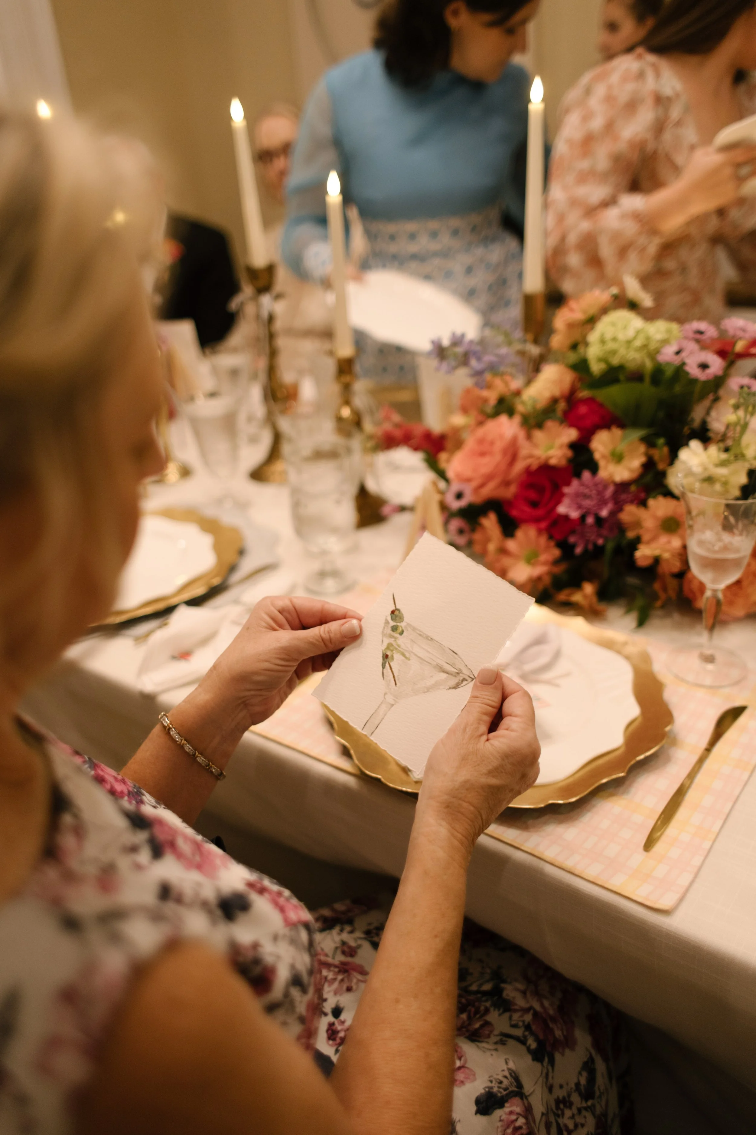 Wedding guest admiring a hand-painted escort card beside colorful floral centerpieces and candlelit reception decor