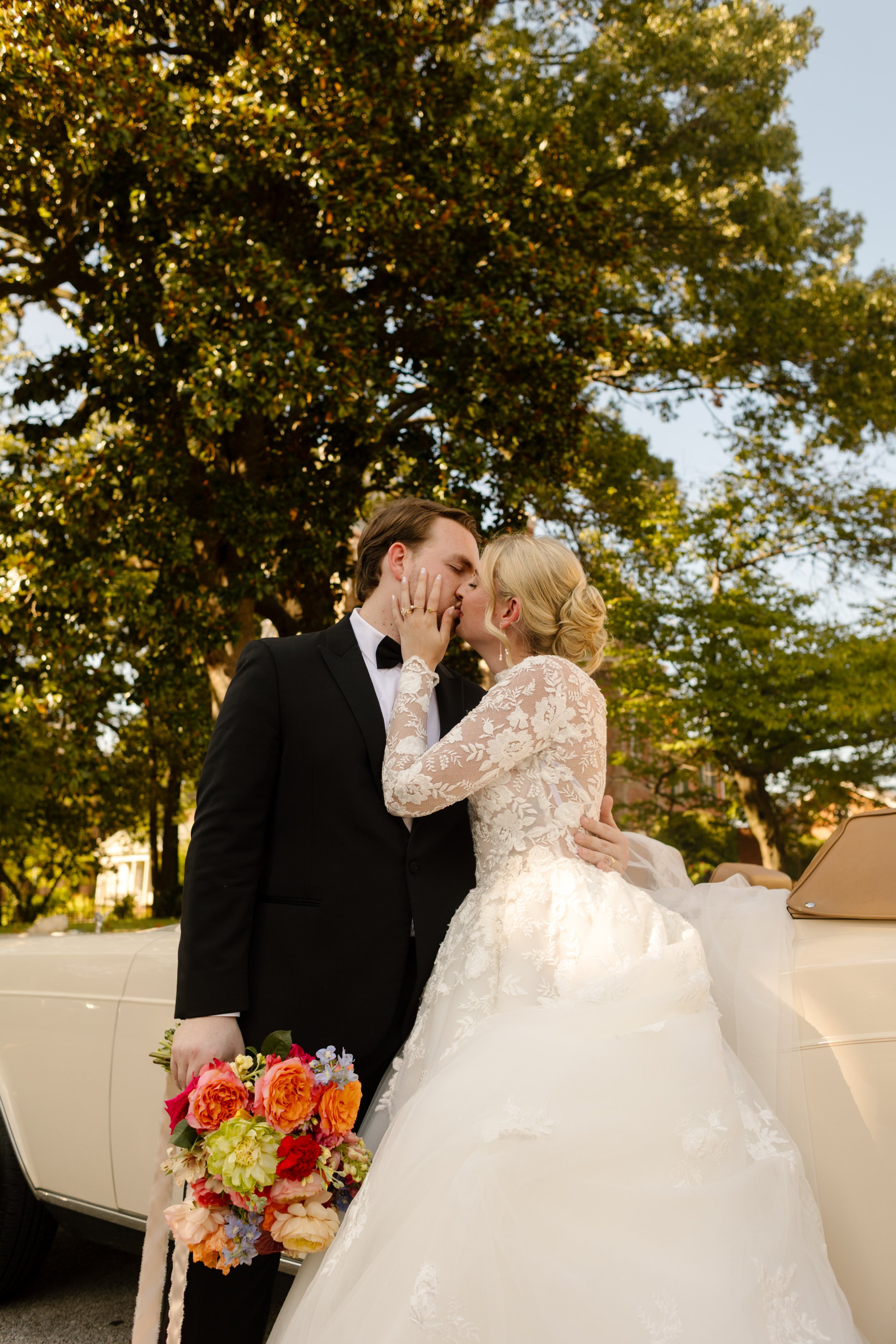 The bride and groom sharing a kiss beside their vintage convertible surrounded by vibrant wedding florals