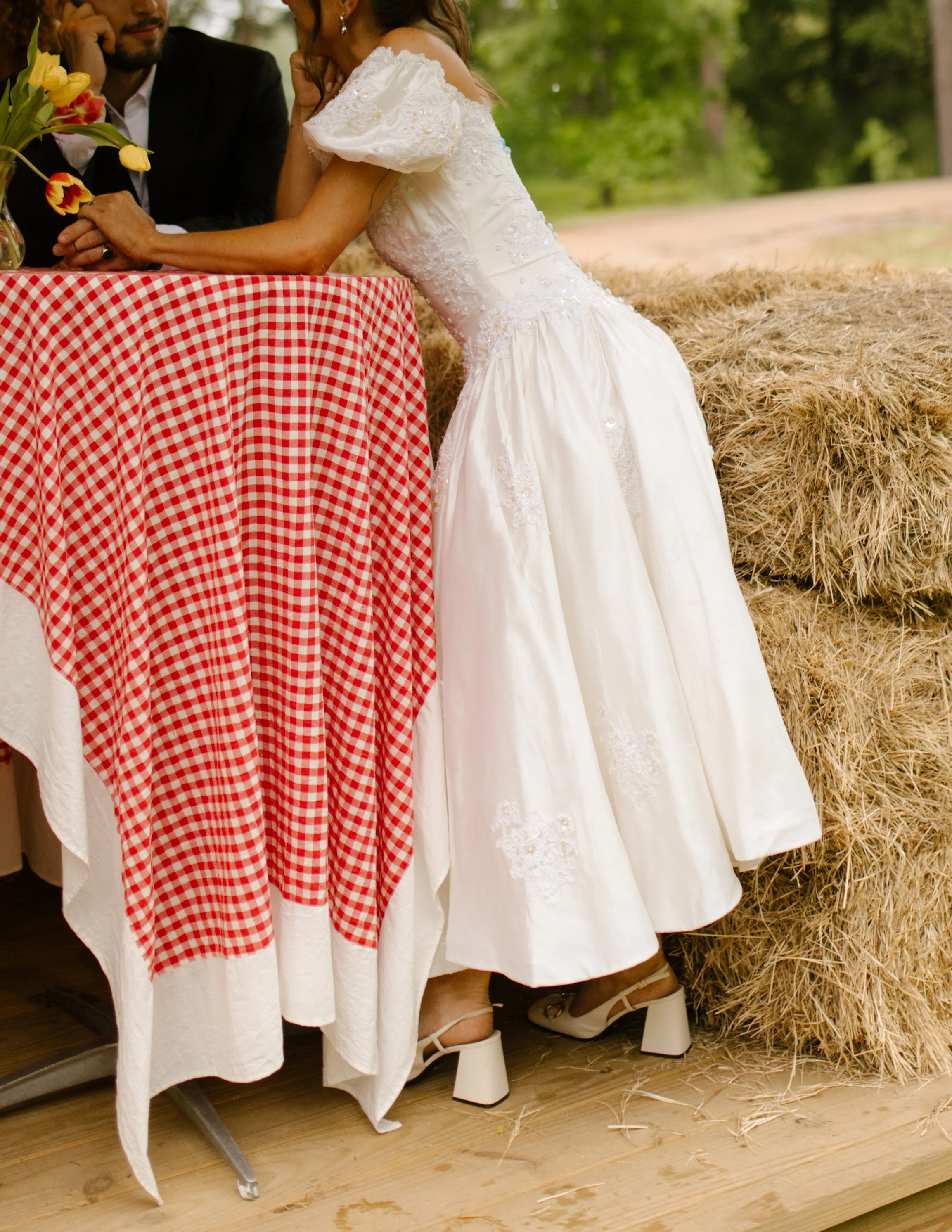 Bride leaning against a gingham-covered cocktail table beside hay bales during rehearsal dinner photography
