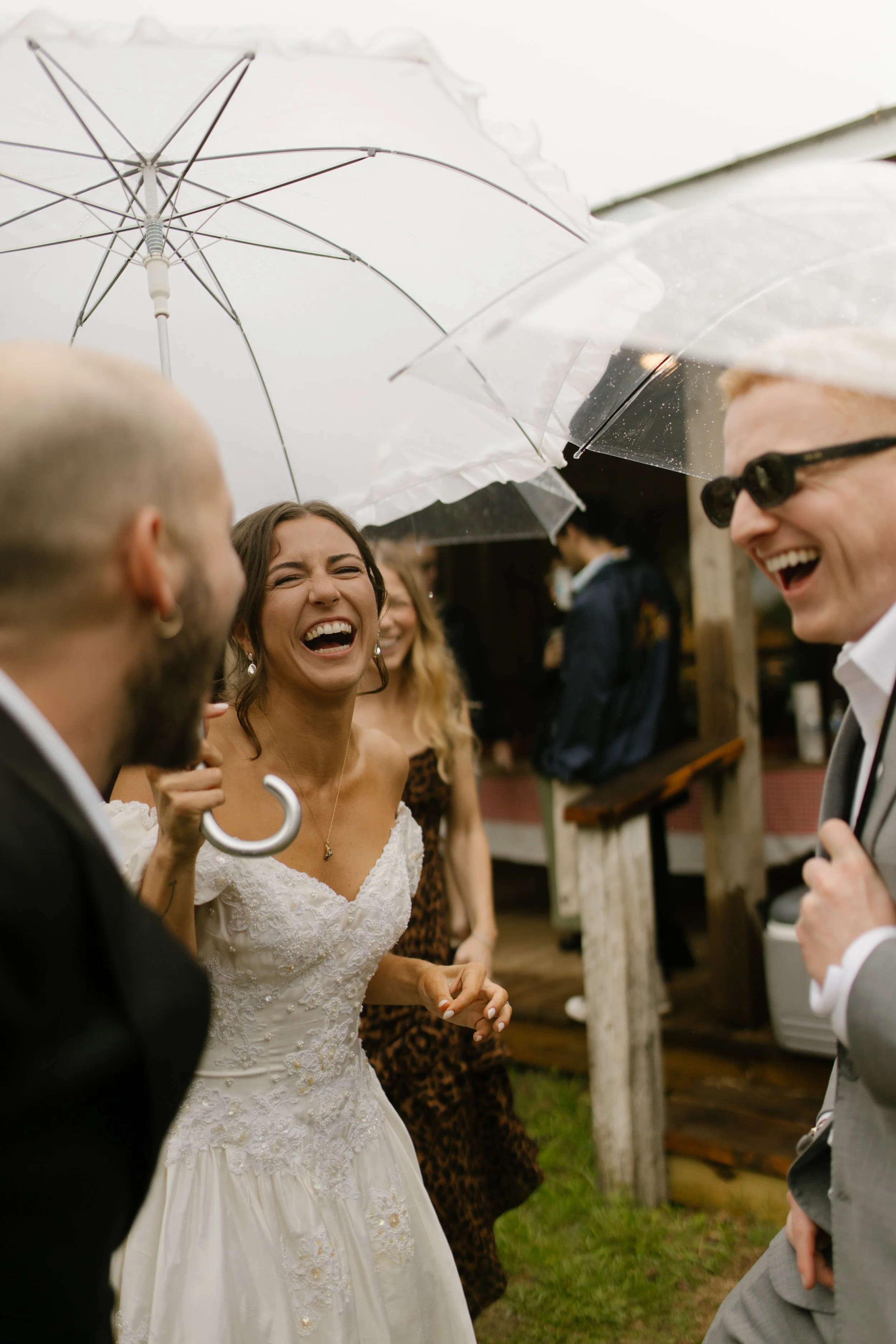 Bride laughing with friends under clear umbrellas during rainy rehearsal dinner photography