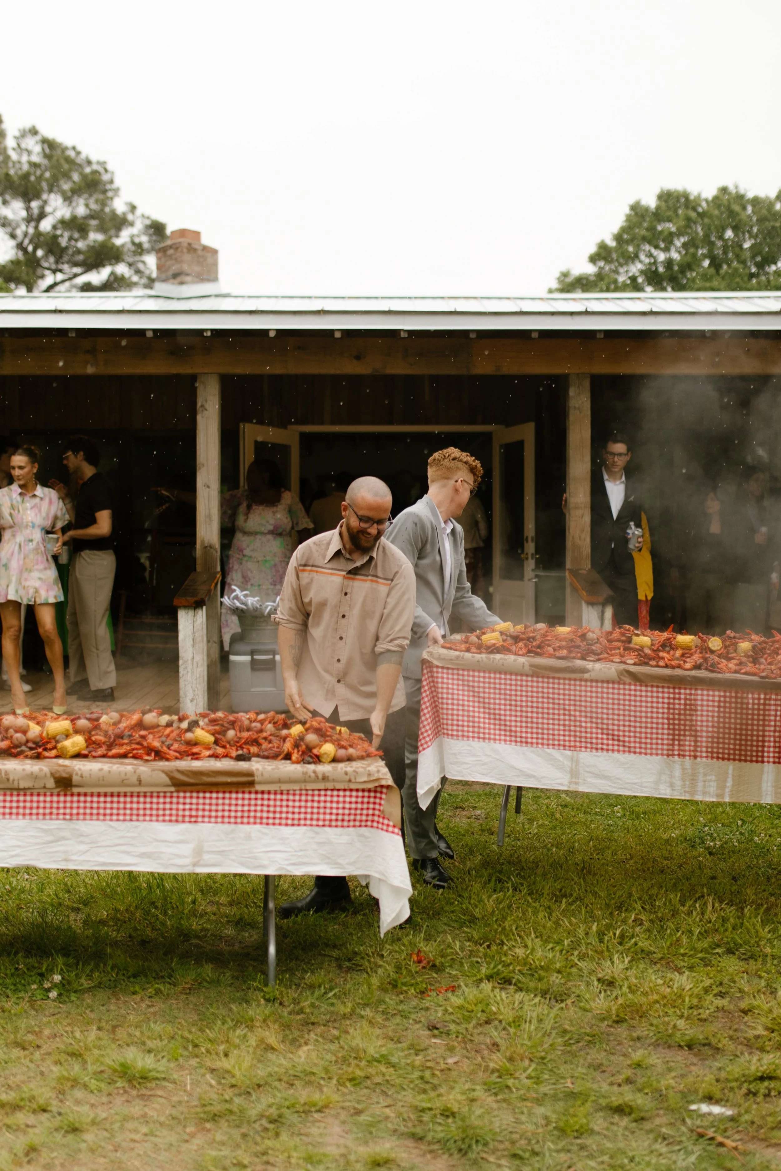 Guests gather around steaming crawfish tables outside a rustic venue during a wedding rehearsal dinner.