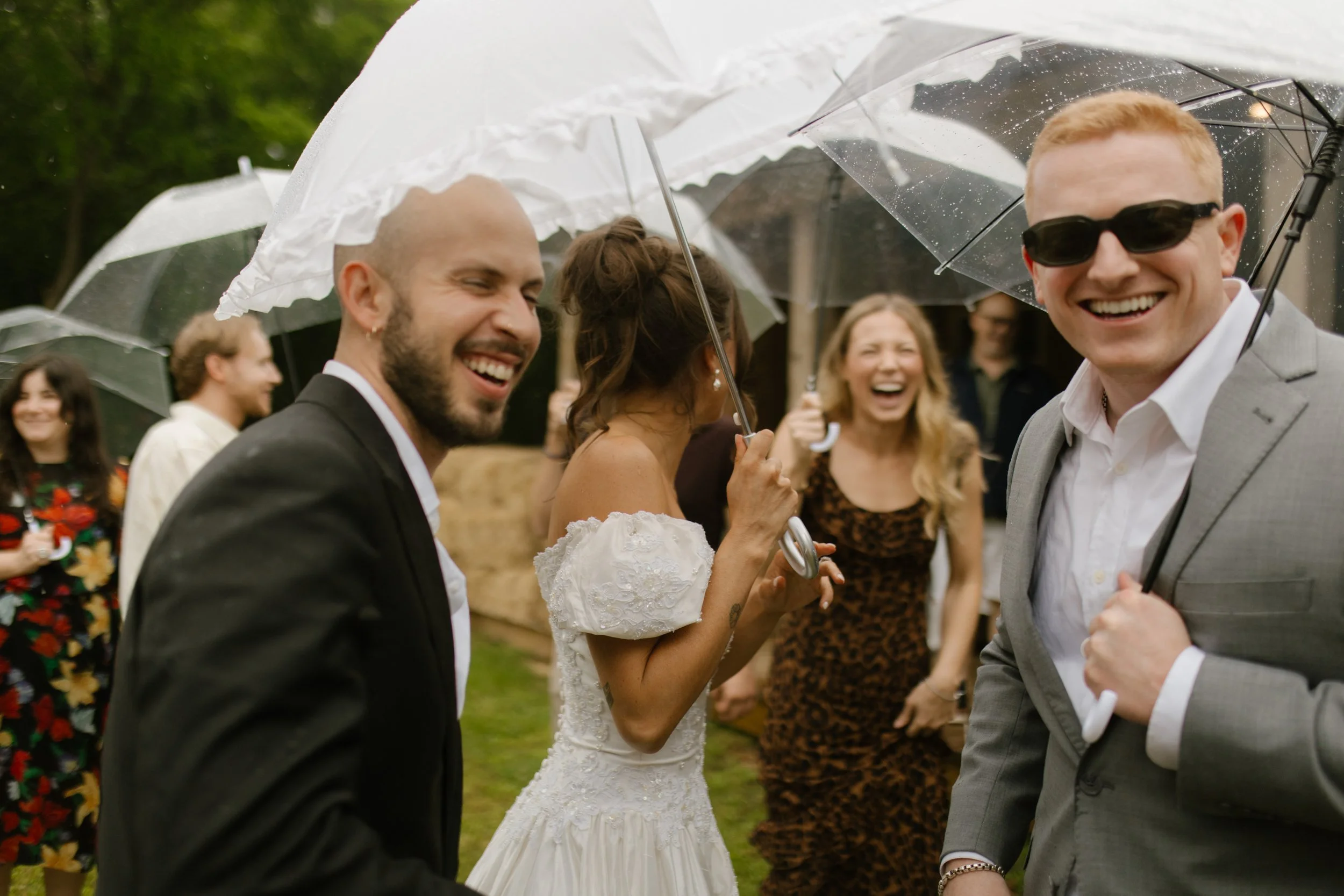 Bride and groom laughing with friends under umbrellas during rainy rehearsal dinner photography