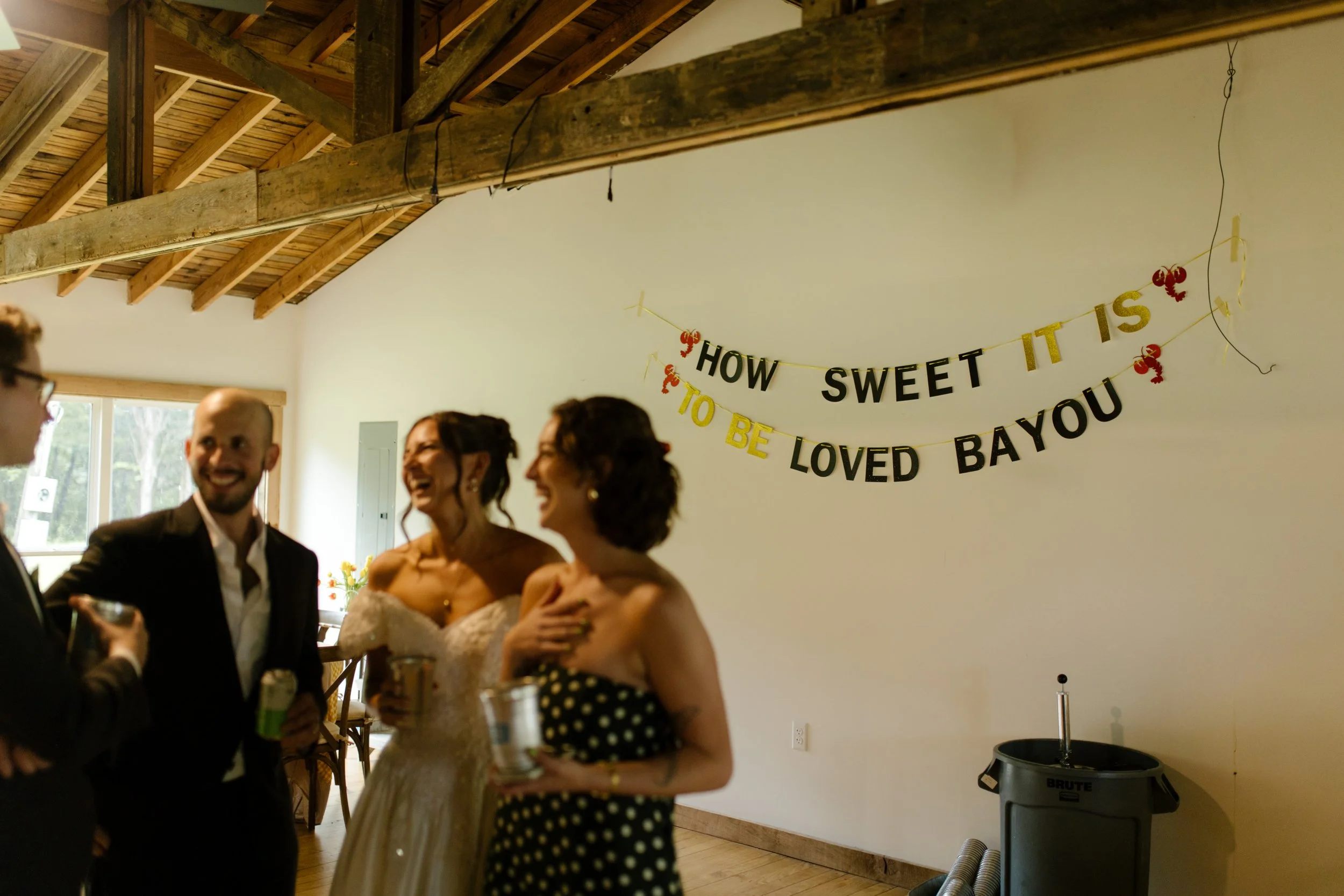 Bride and guests laughing beneath a "How Sweet It Is to Be Loved Bayou" sign during a wedding rehearsal dinner
