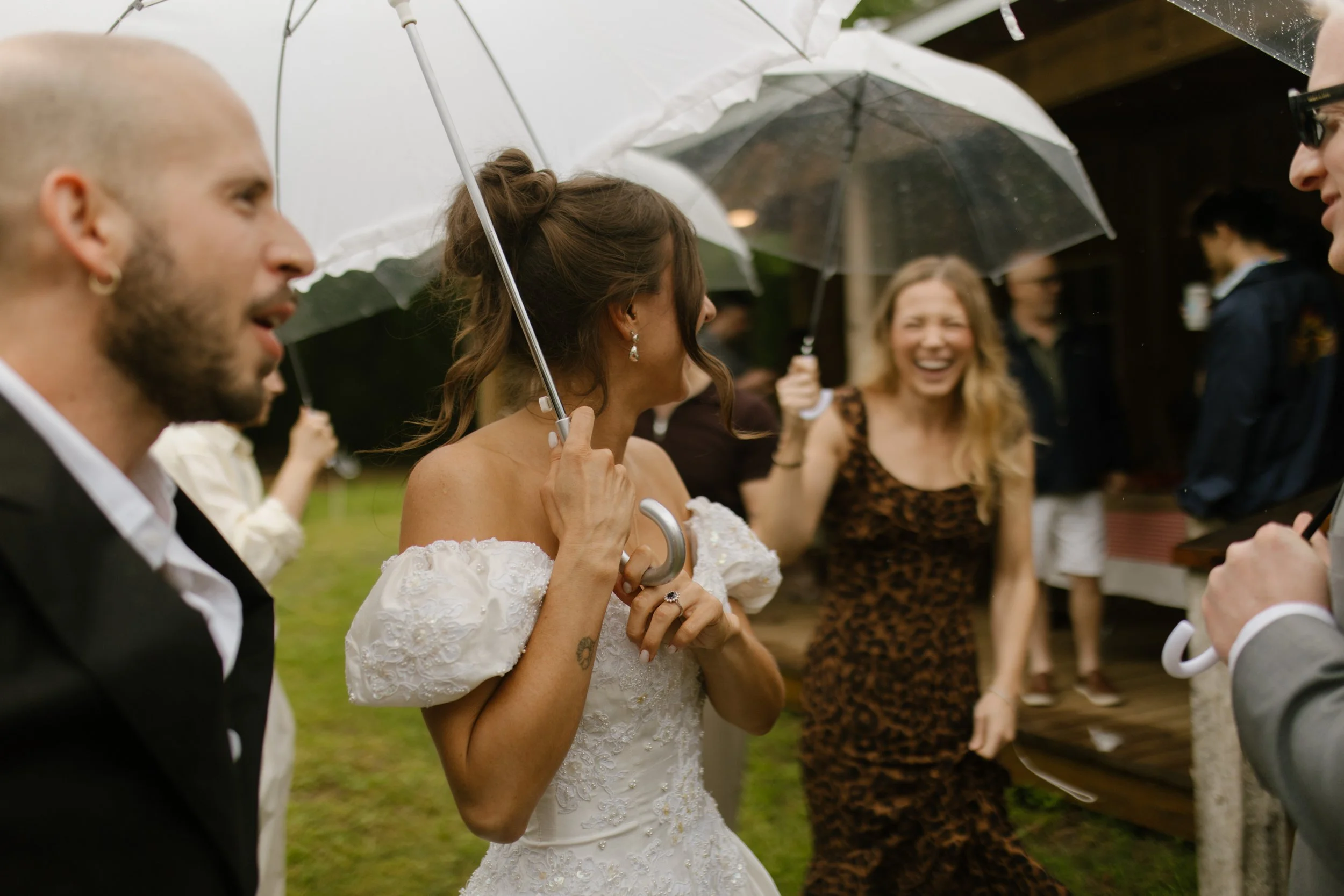 Bride laughing with guests beneath umbrellas while enjoying a rainy outdoor rehearsal event.