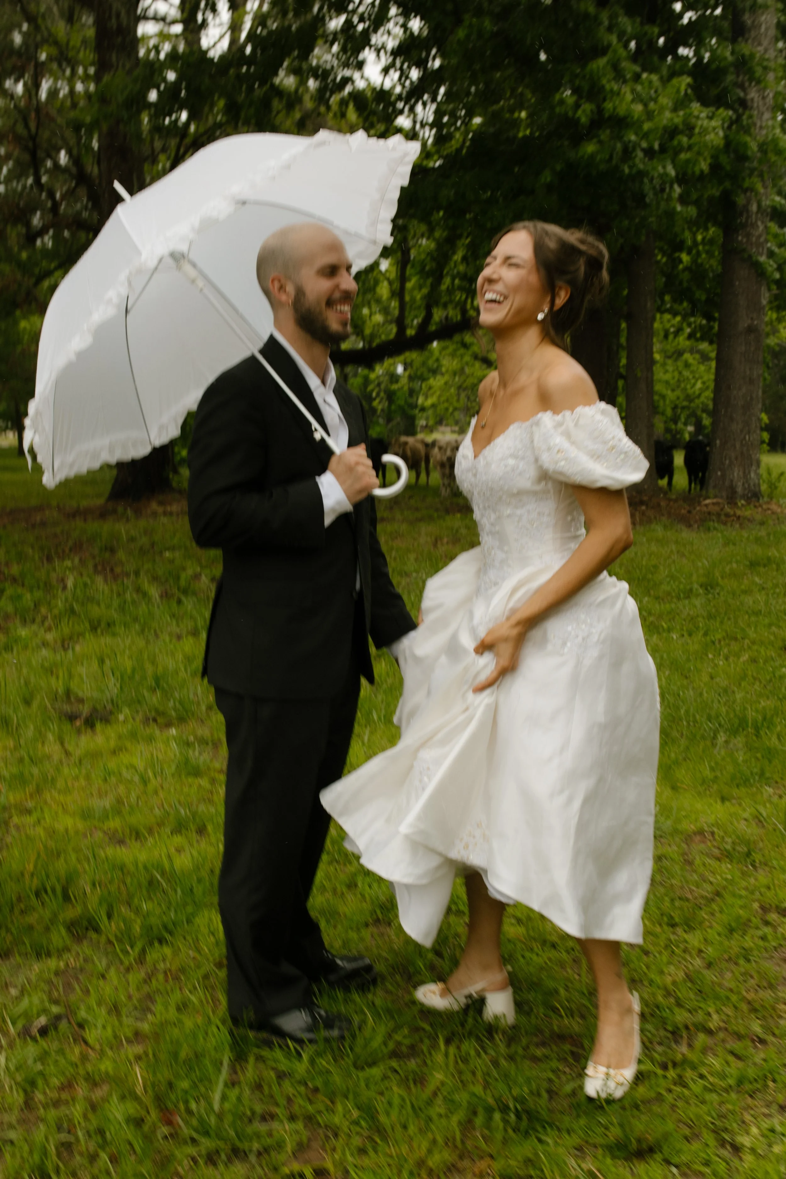Bride and groom laughing together beneath an umbrella in a rain-soaked field during wedding rehearsal photography.