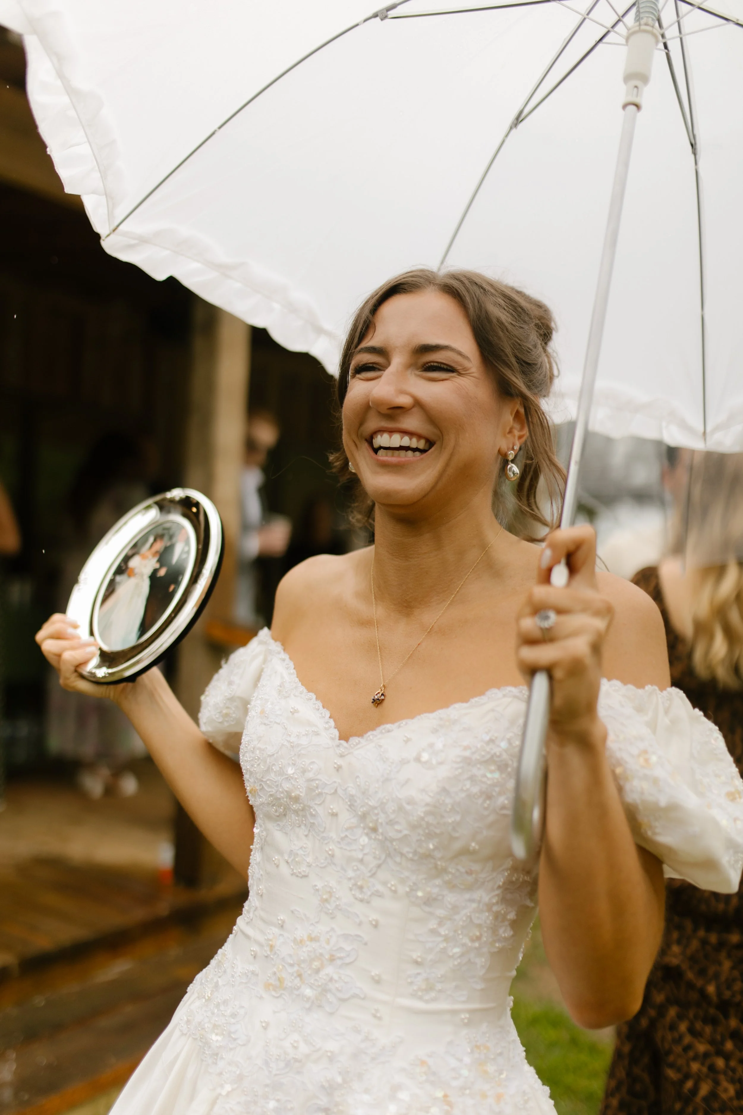 Bride smiling while holding an umbrella and framed photo during a rainy rehearsal dinner celebration.