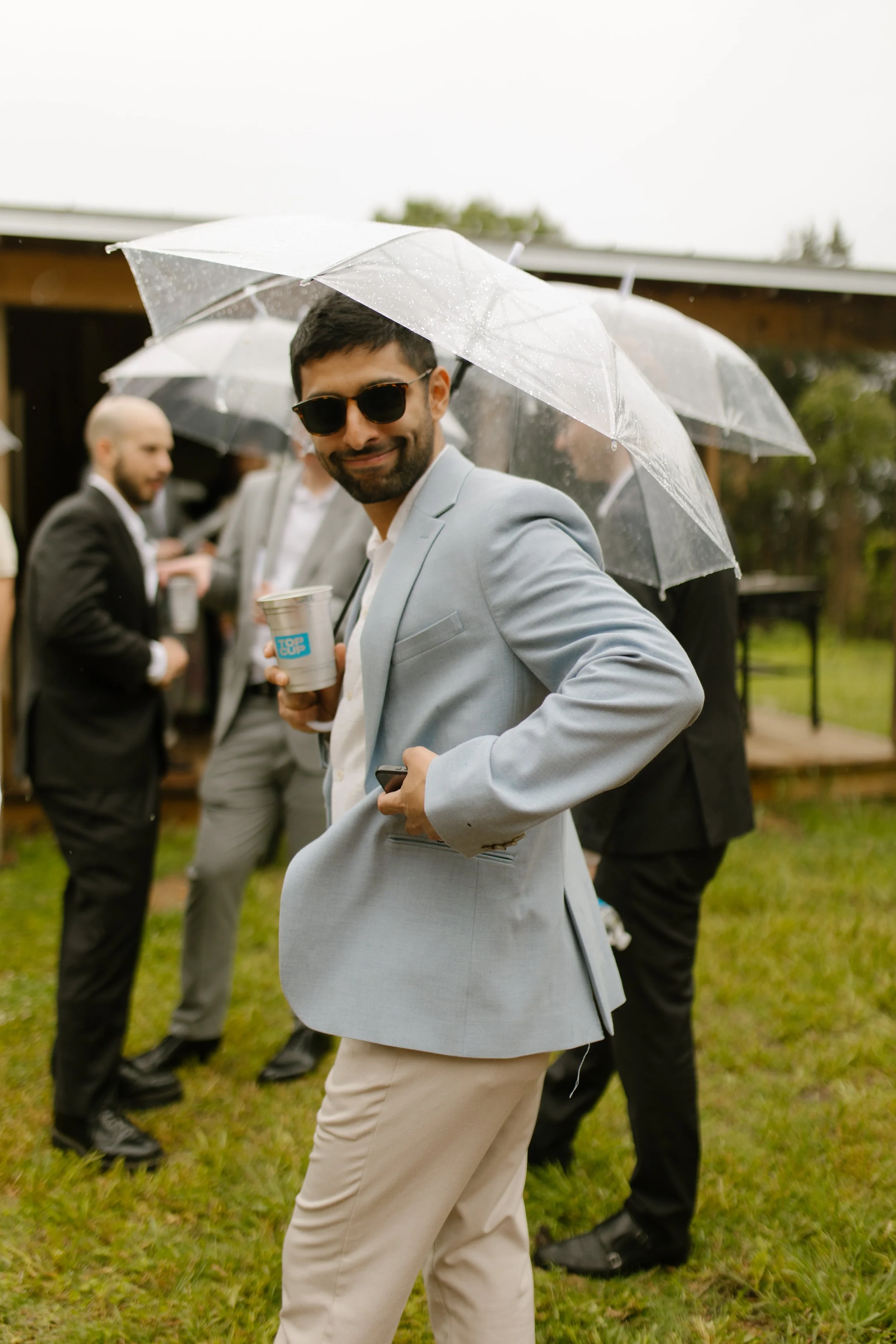 Guest in a light blue blazer posing with a drink under a clear umbrella at the rehearsal gathering.